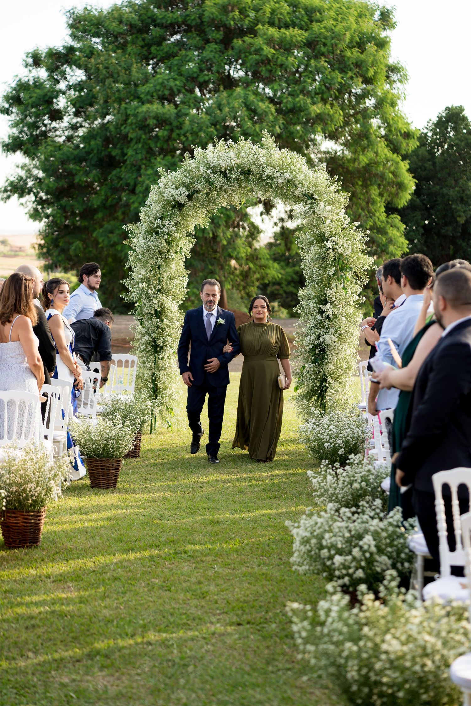 Couple walks through white floral arch at outdoor wedding ceremony with guests seated on either side.