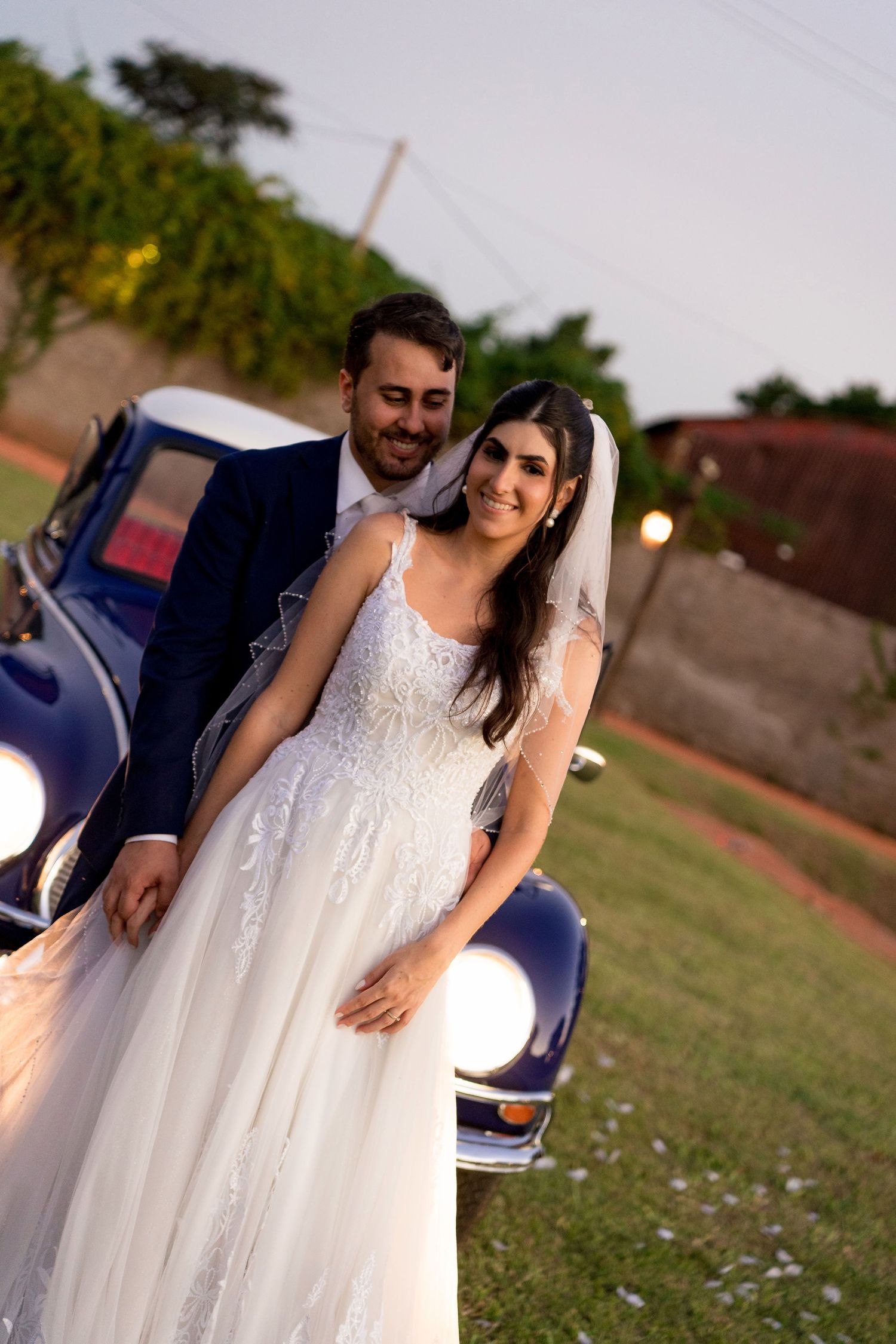 A smiling bride in a white lace gown and groom in navy suit pose together beside a vintage blue car at dusk.