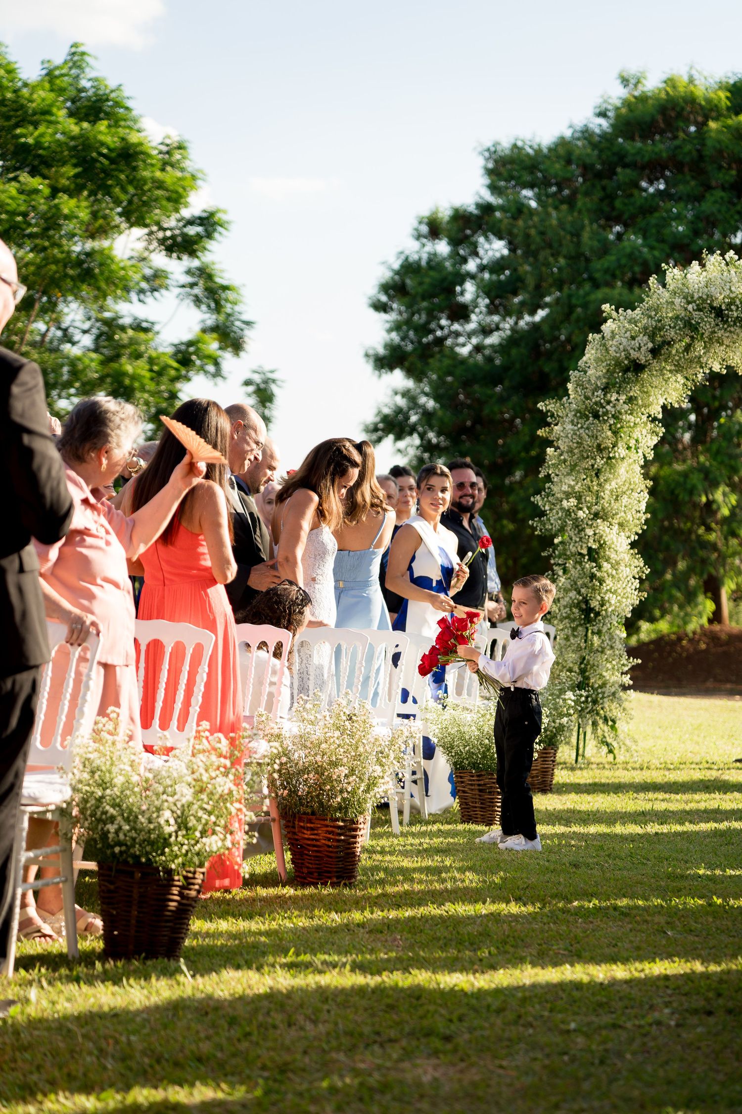 Young ring bearer walks down outdoor wedding aisle flanked by guests and floral arch on sunny day.