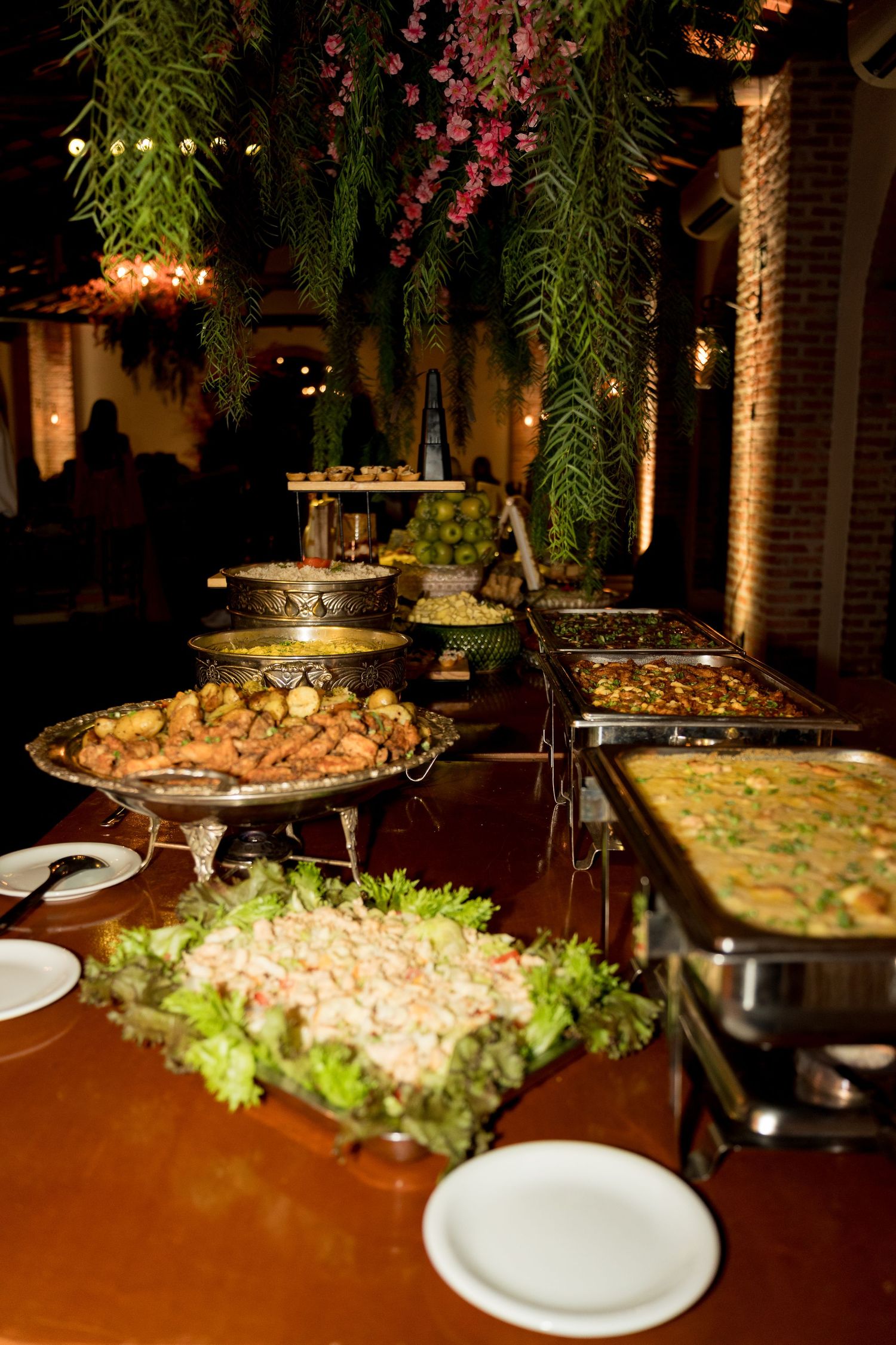 Elegant buffet table with assorted dishes, chafing trays, and lush floral decor at an indoor evening event.