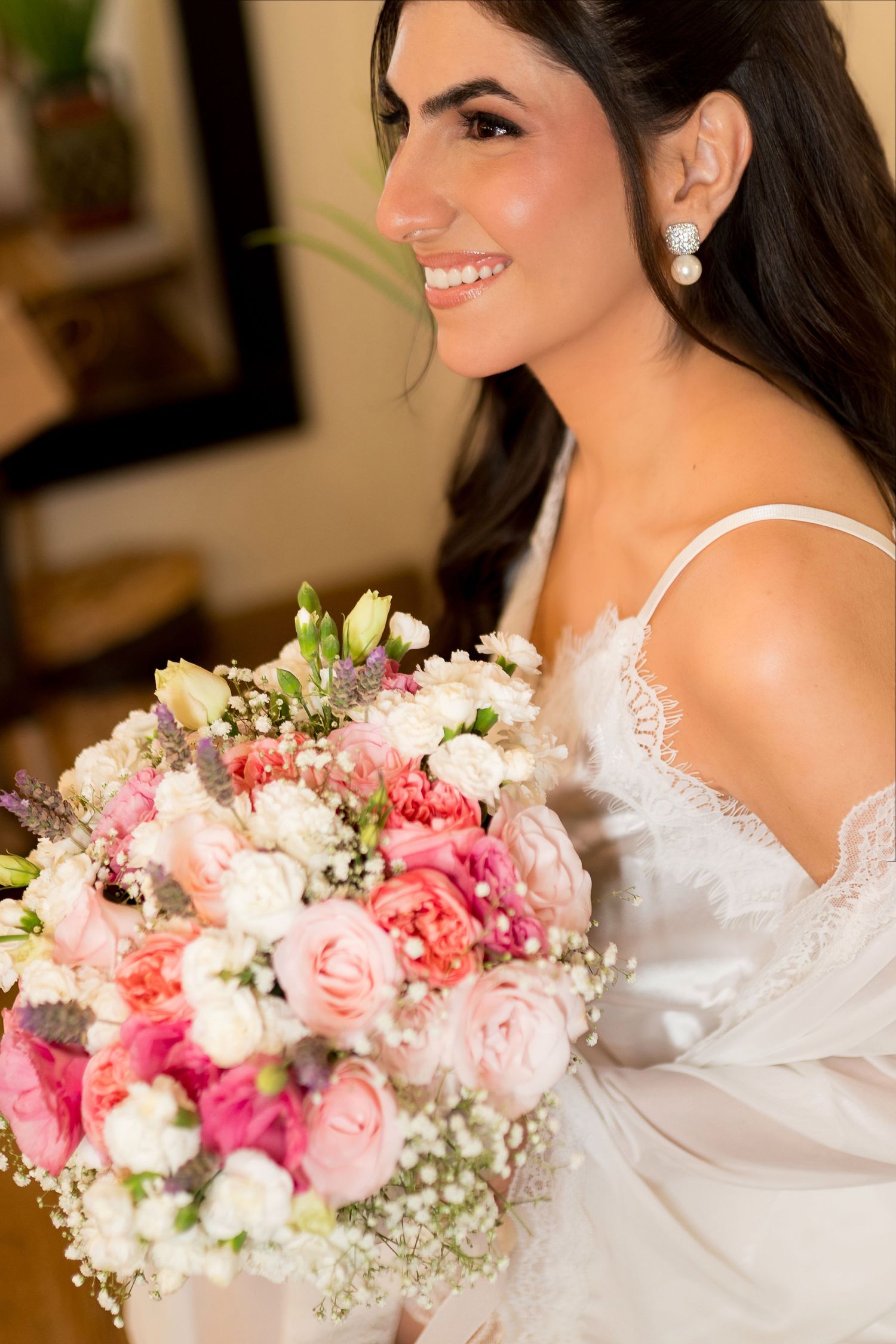 Smiling bride in white lace gown holding a lush pink and white floral bouquet with roses and baby's breath.