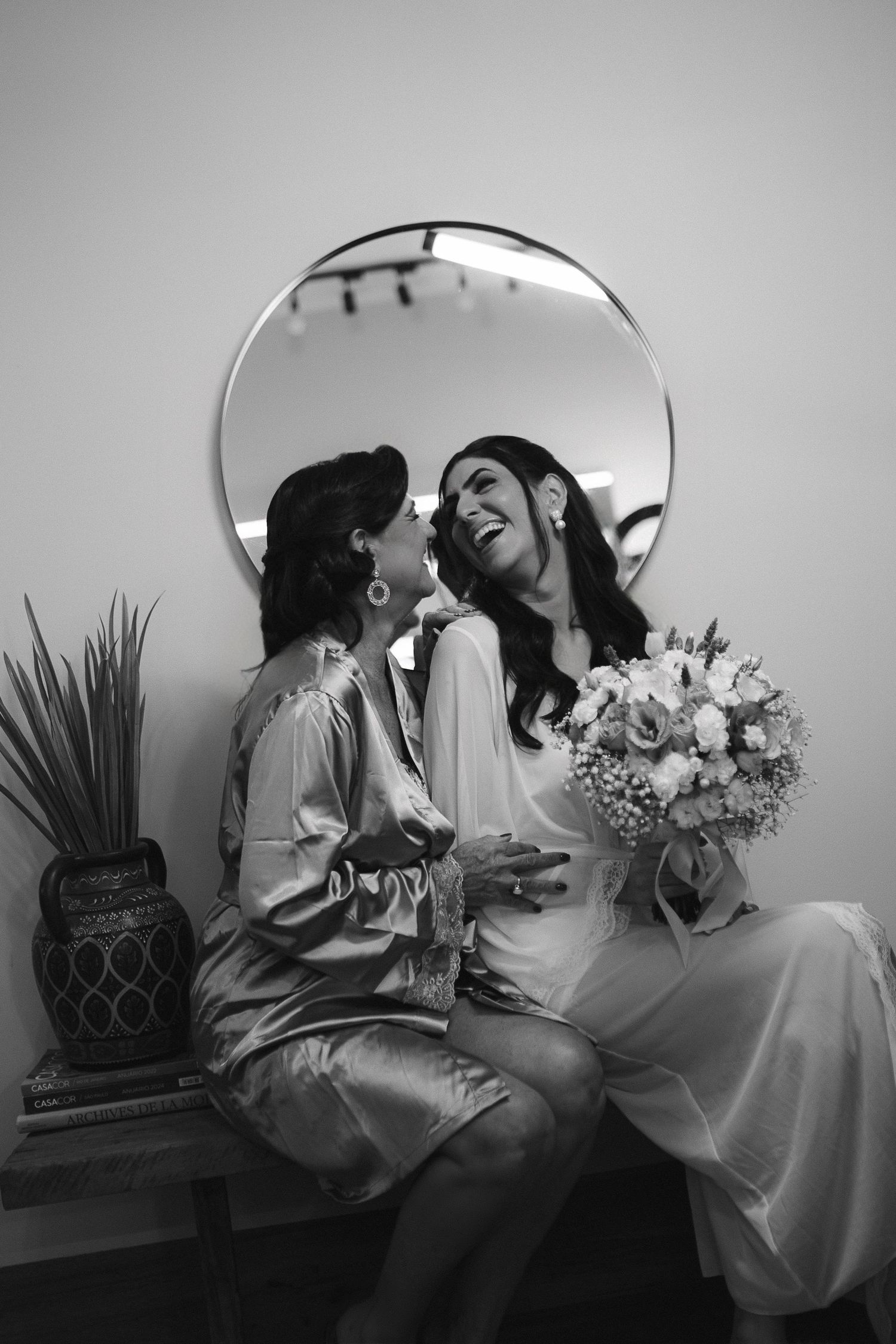 Black and white photo of two women laughing together, one in a wedding dress holding a bouquet, seated before a round mirror.