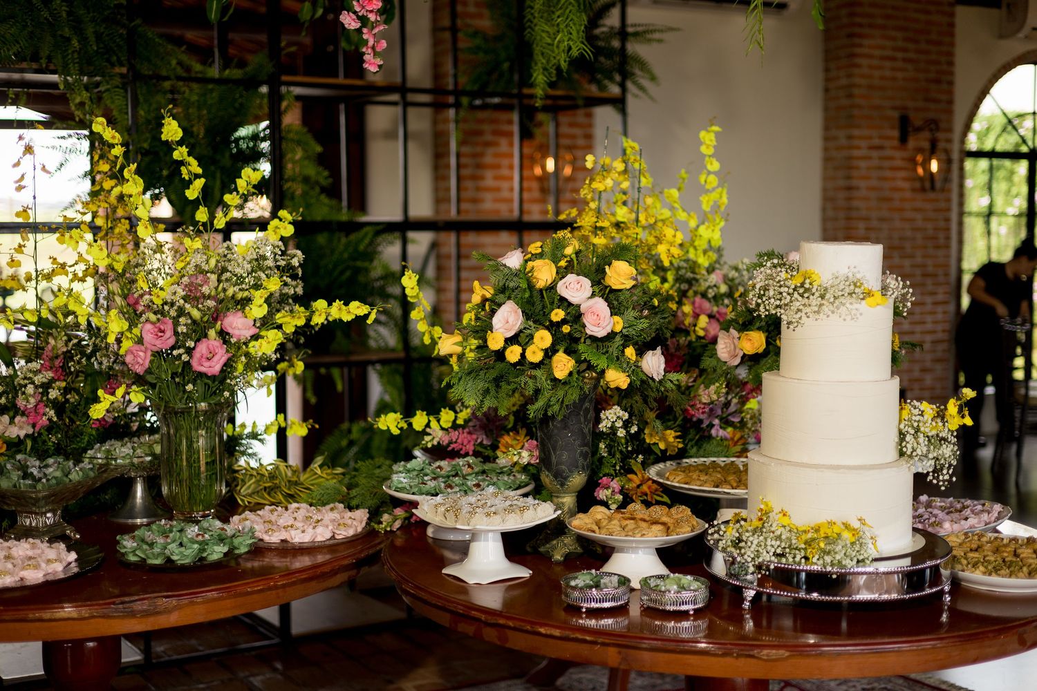 Elegant wedding dessert table with tiered white cake, colorful floral arrangements in yellow and pink, and assorted sweets.