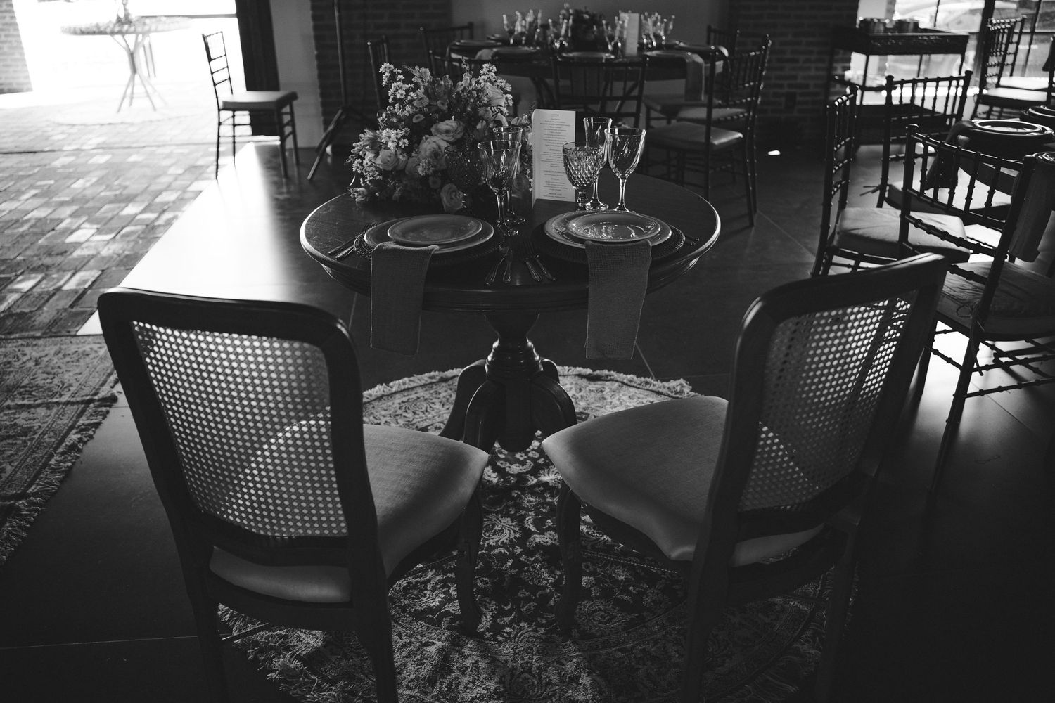 Black and white photo of an elegant dining table setup with cane chairs and floral centerpiece on a patterned rug.