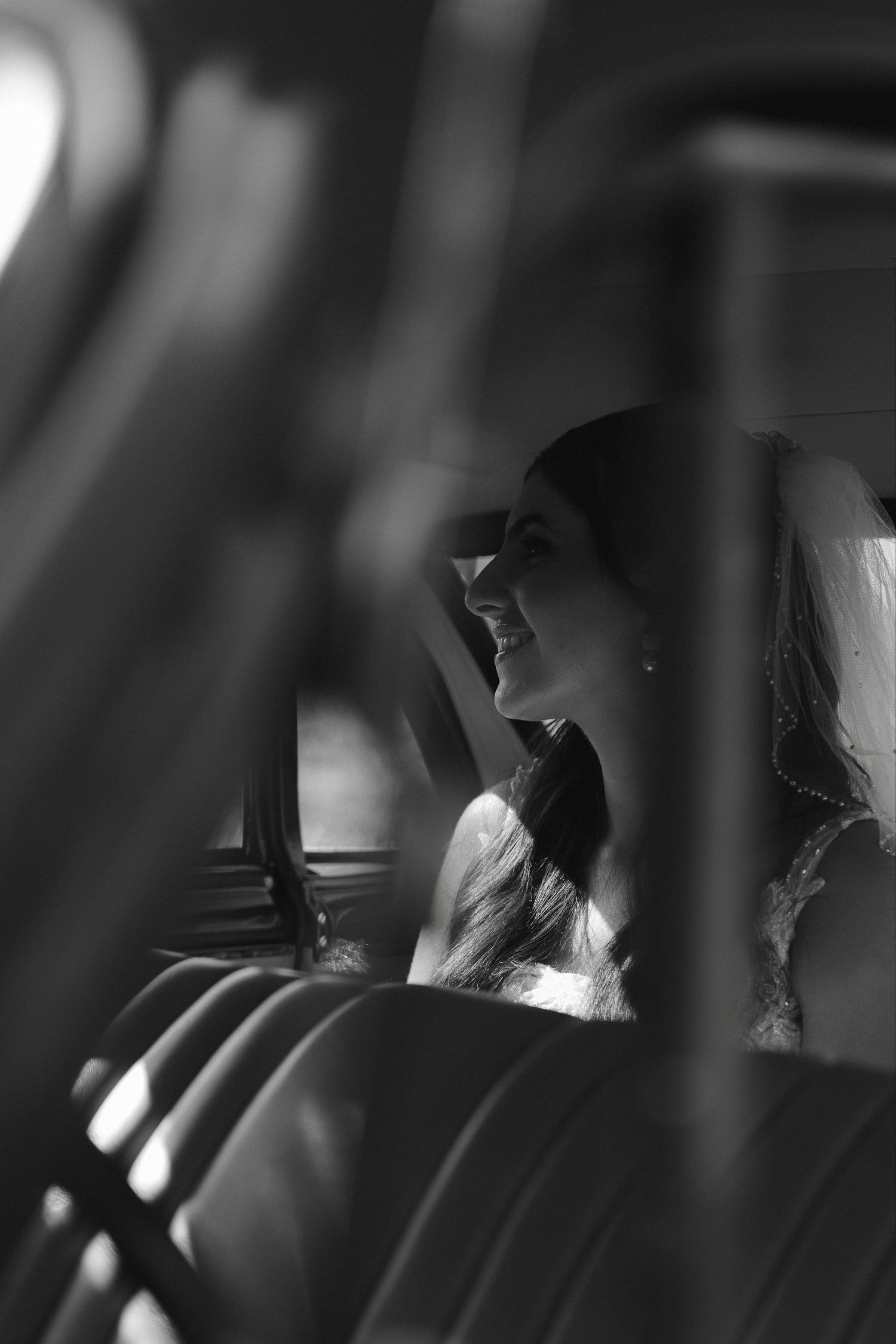 Black and white photo of a smiling bride in a veil sitting in a vintage car, sunlight streaming through the window.