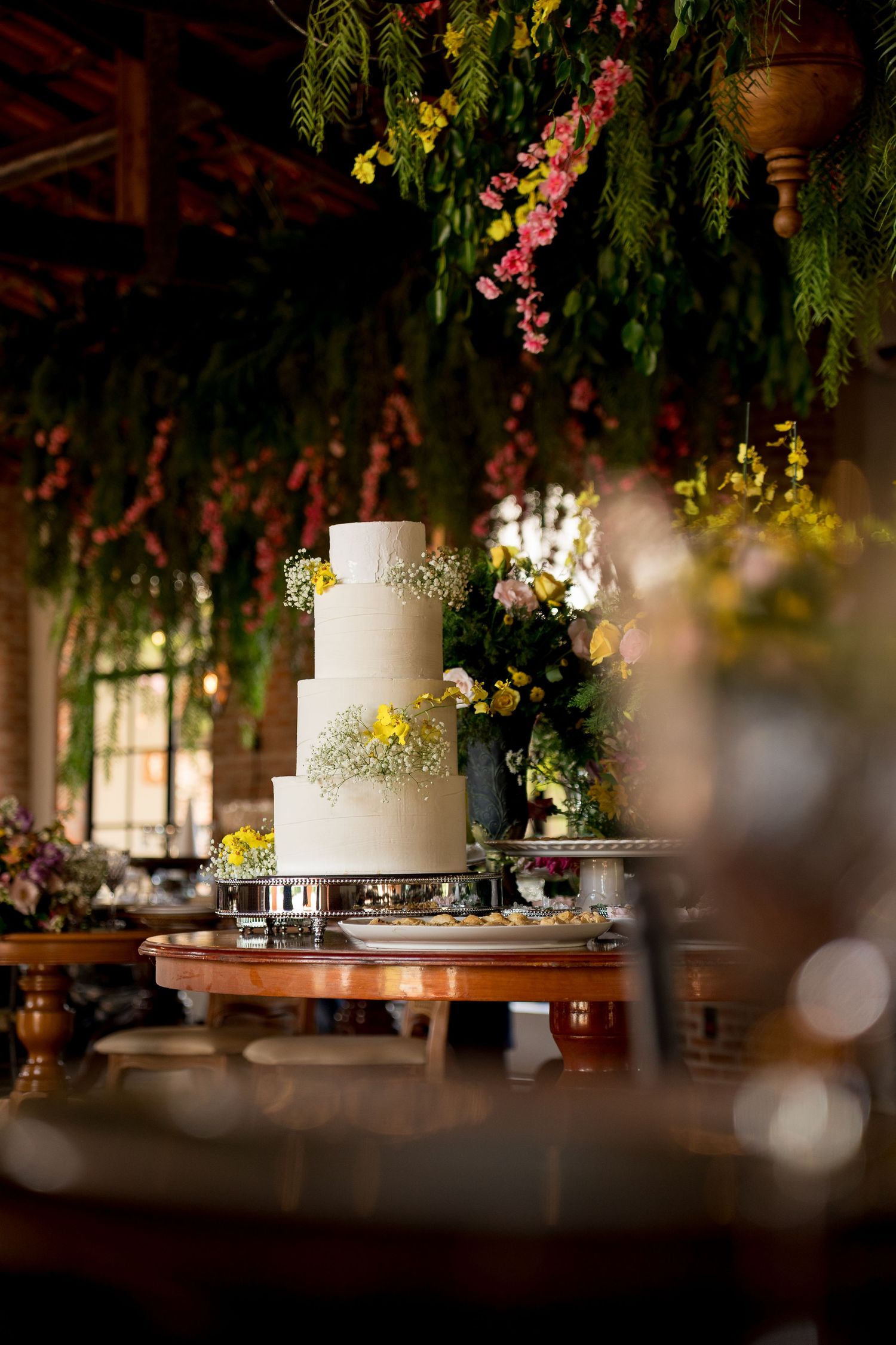 Elegant three-tier white wedding cake adorned with yellow flowers displayed on a silver stand beneath lush floral ceiling decor.