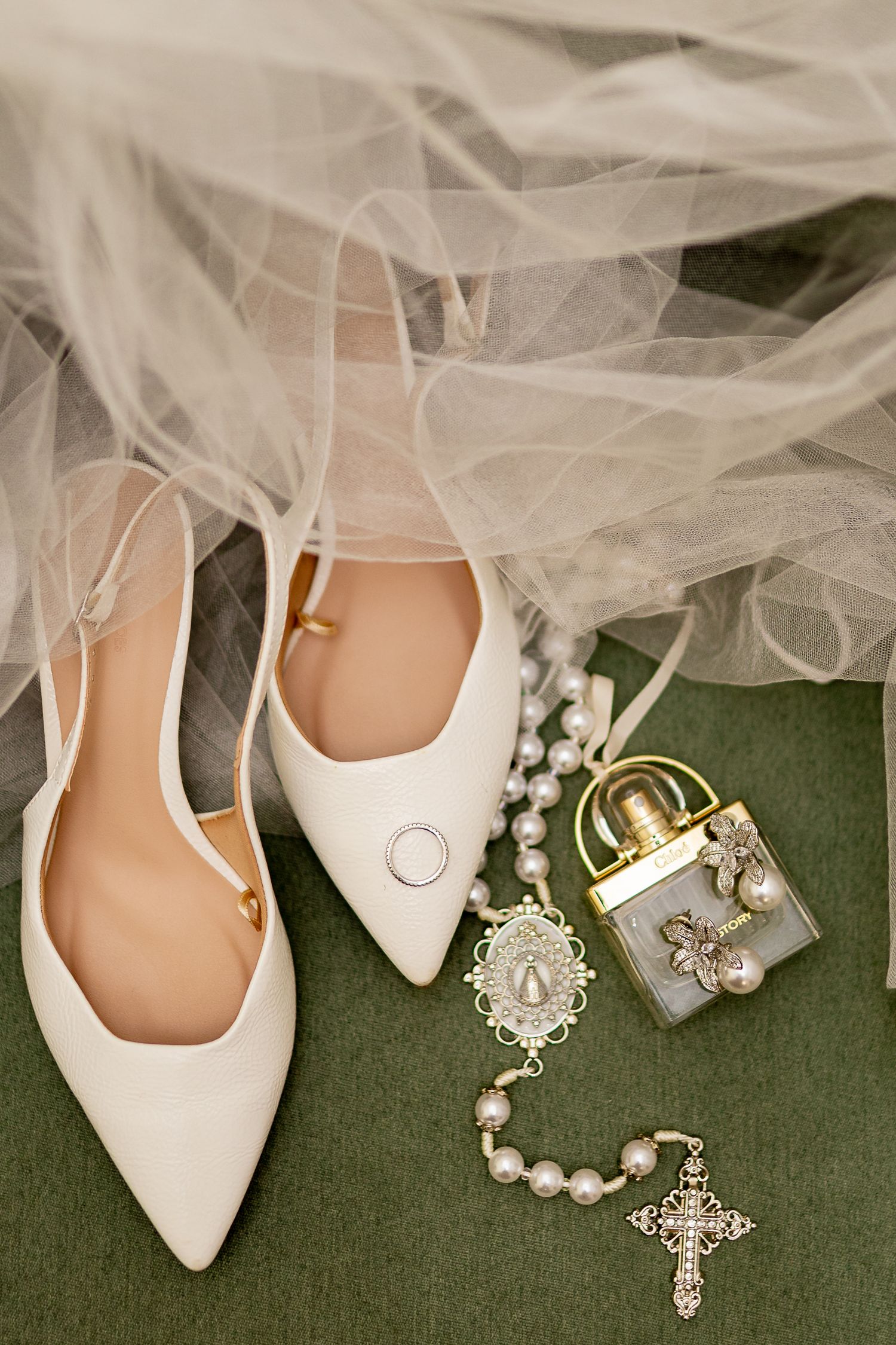 White bridal heels, pearl rosary, ring, and gold perfume bottle arranged on green surface with tulle veil.