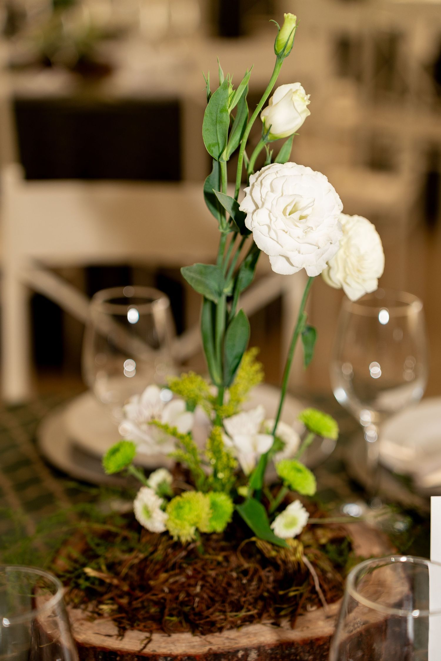 Elegant white ranunculus and lisianthus floral centerpiece with green accents on a moss base at a formal table setting.