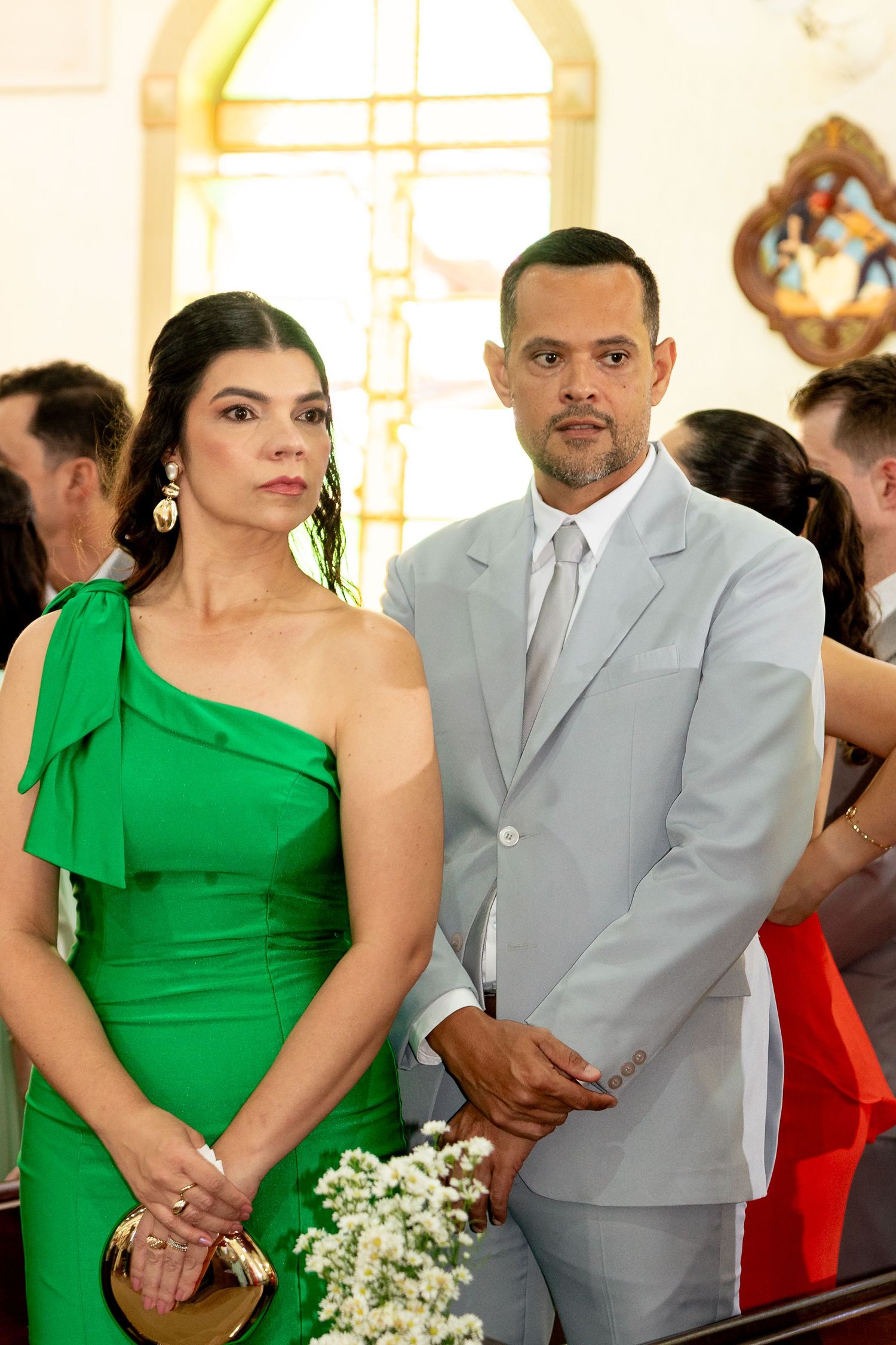 Elegant couple in a church, woman in green dress and man in light gray suit, attending a formal wedding ceremony.