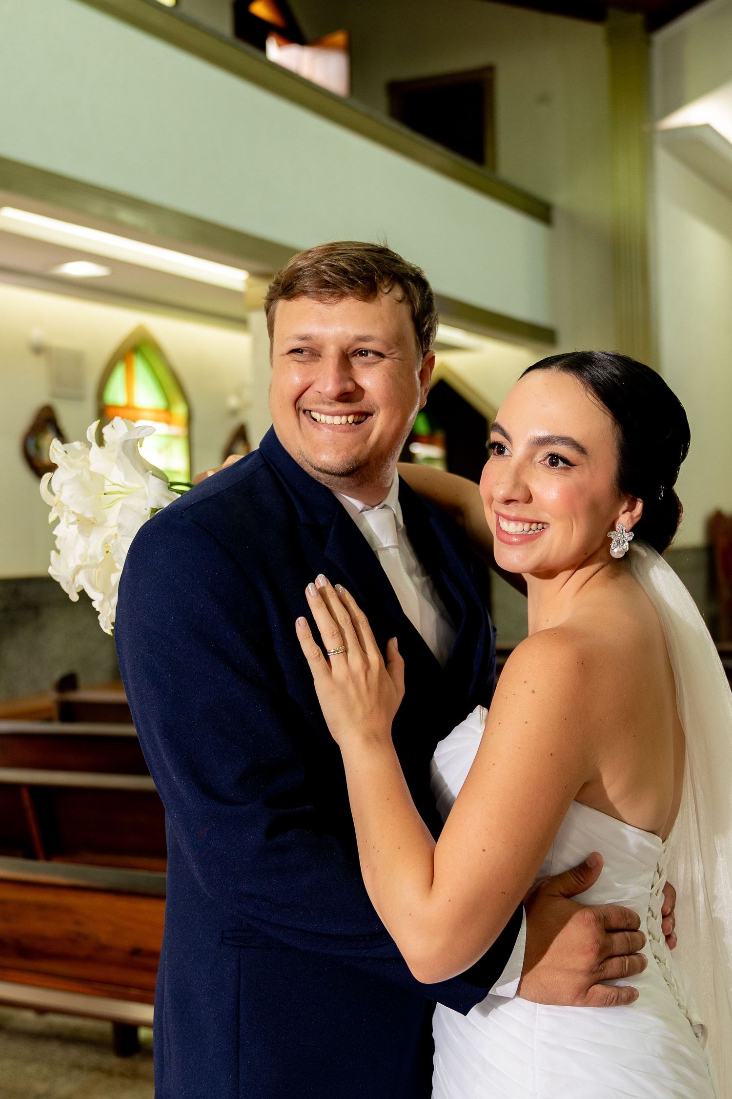 Happy newlyweds pose together in a church, bride in white gown with veil, groom in navy suit holding white flowers.