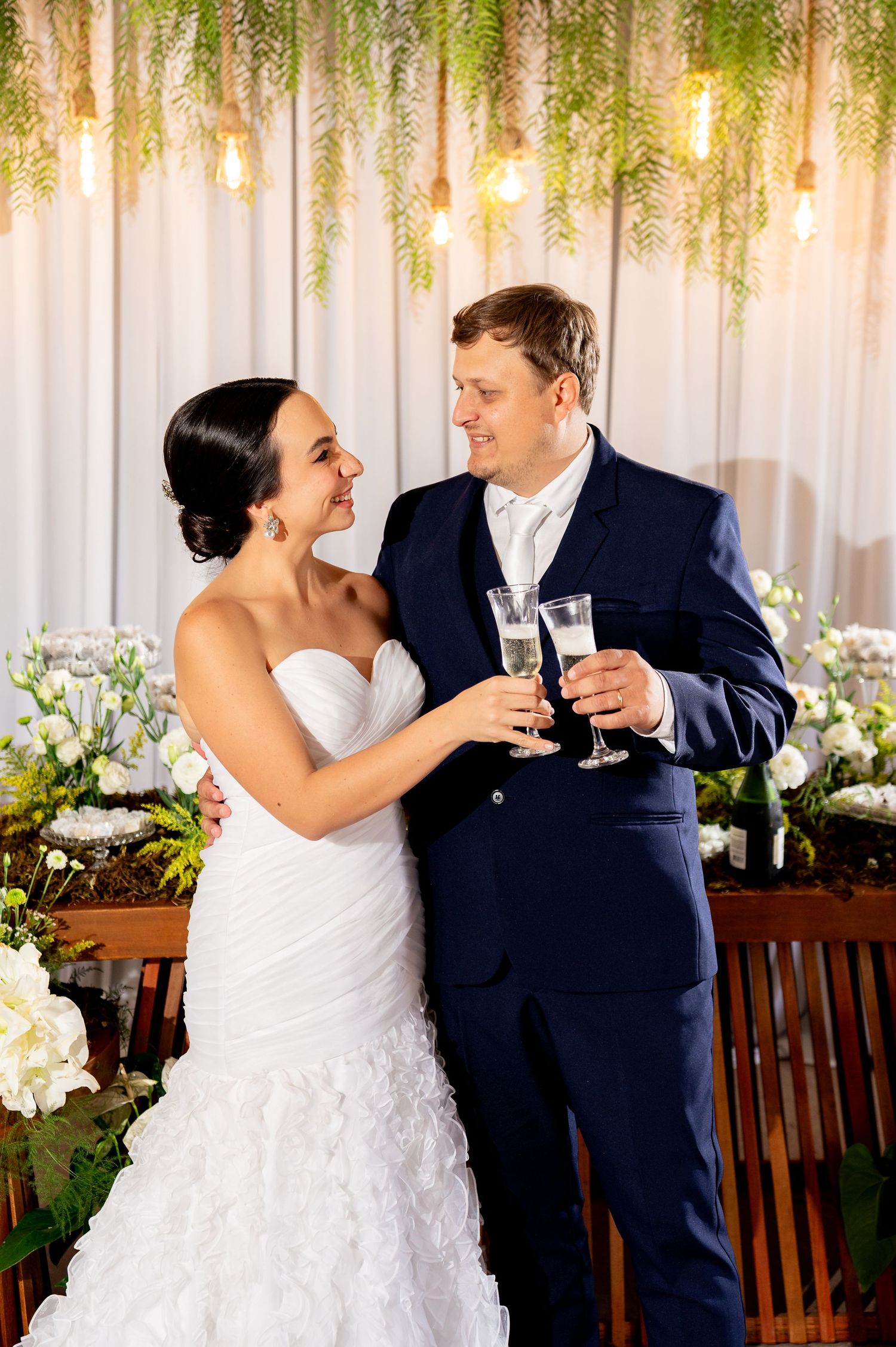 Bride and groom toasting with champagne glasses at wedding reception, surrounded by floral decor and hanging greenery.