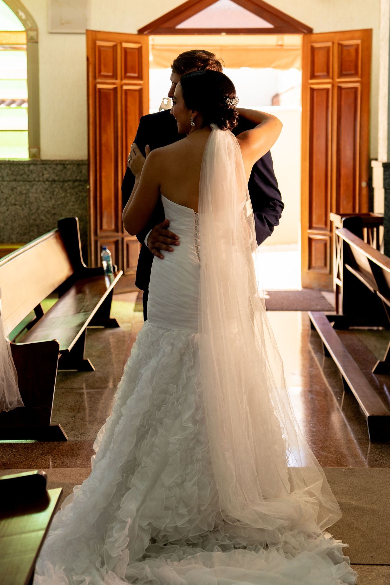 Bride and groom embracing at church altar, bride in elegant white mermaid gown with long veil, viewed from behind.