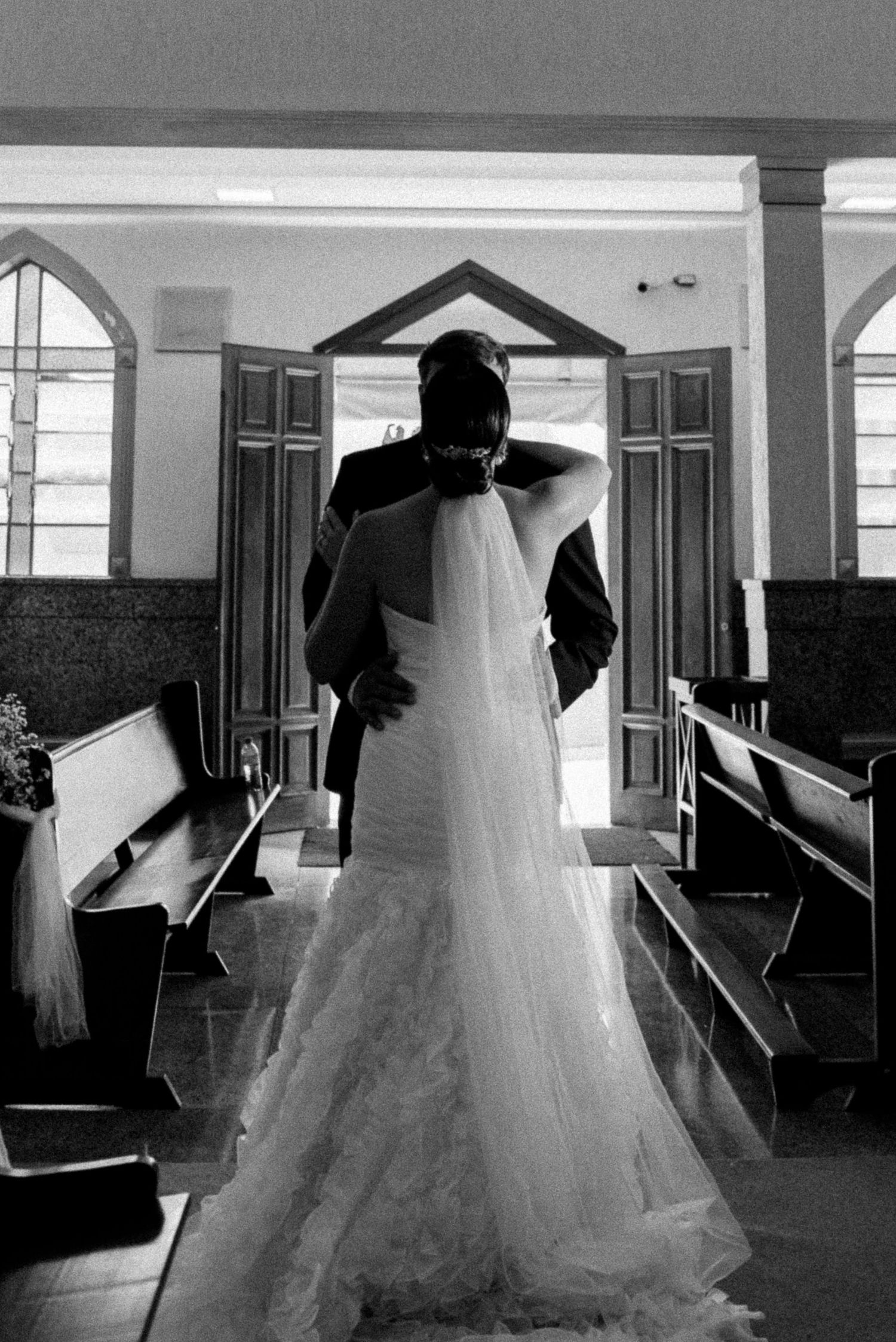Black and white photo of bride and groom embracing at church altar, wedding dress with veil, wooden pews in background.