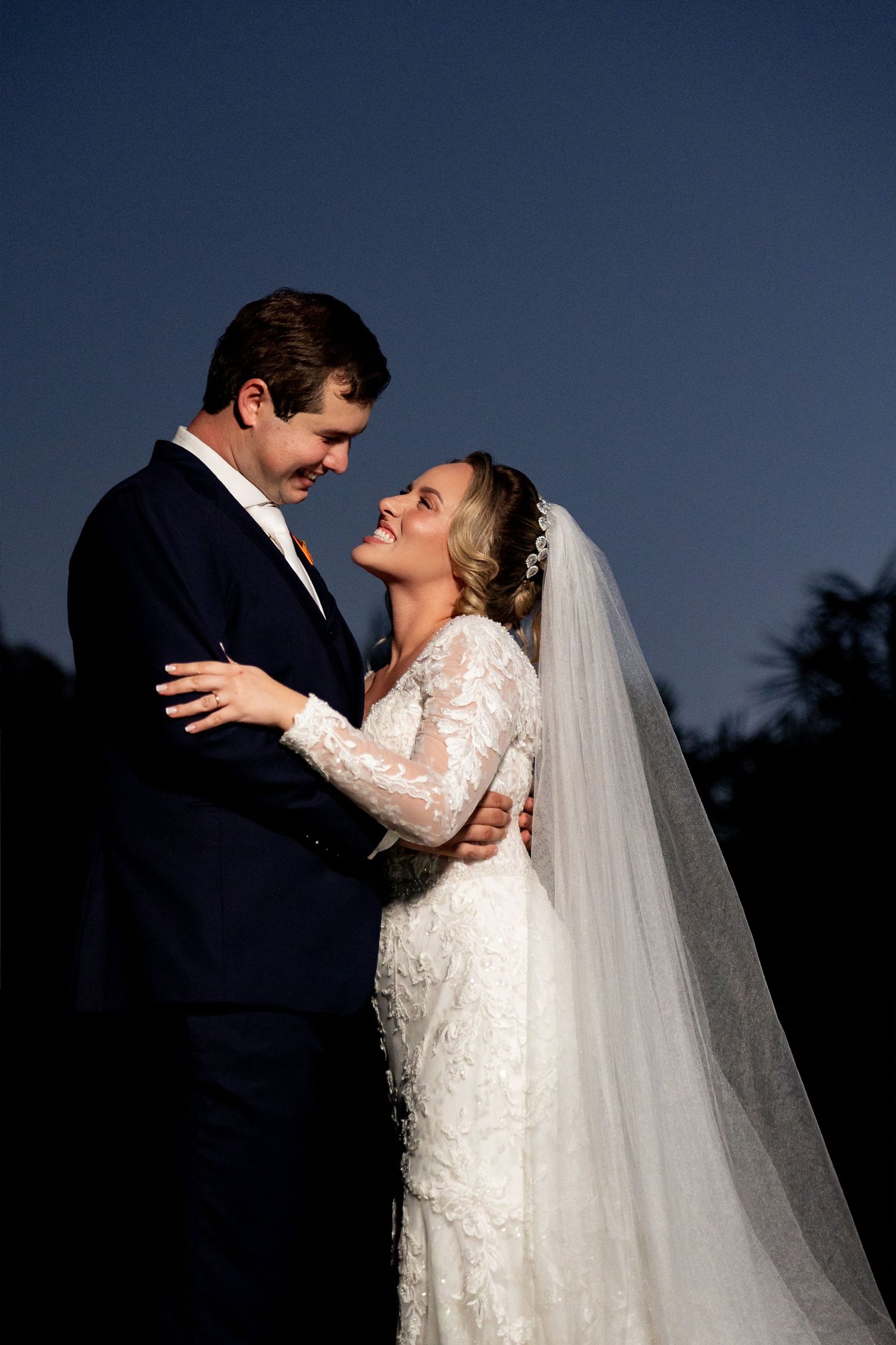 Bride and groom share a romantic embrace at dusk, with bride in lace gown and veil smiling up at her groom.
