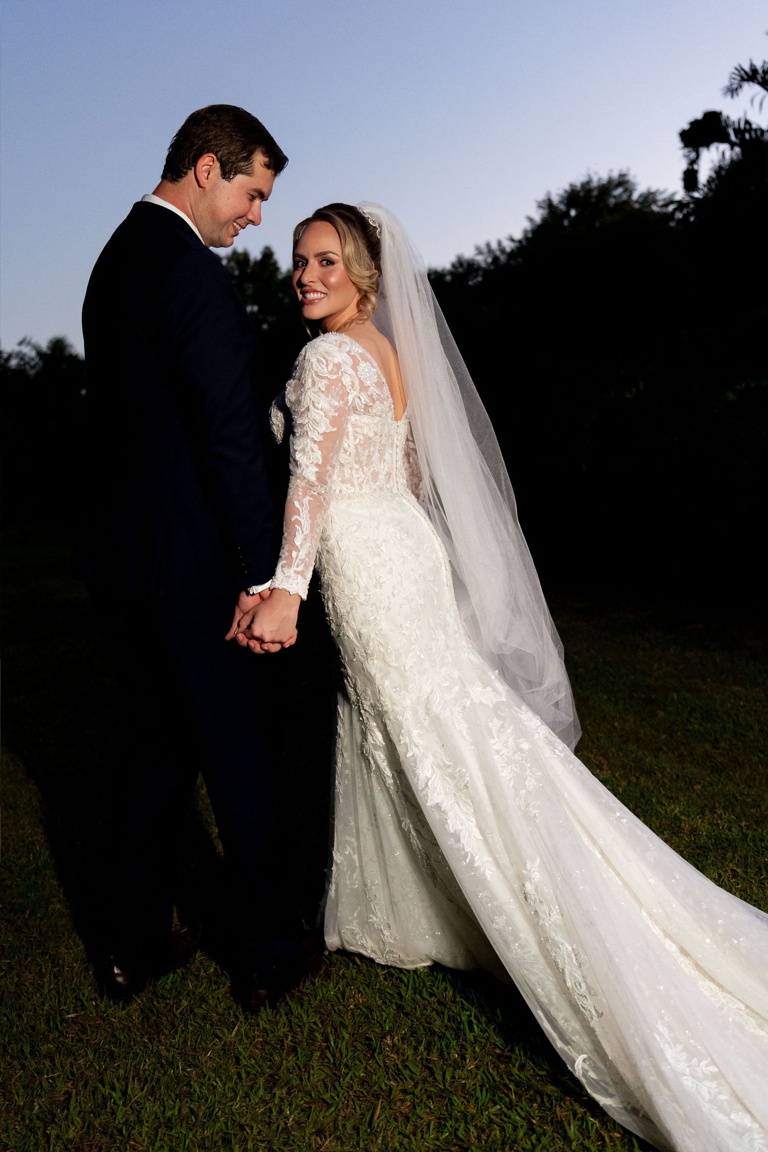 Bride in lace long-sleeve wedding gown with veil and groom in black tuxedo holding hands outdoors at dusk.