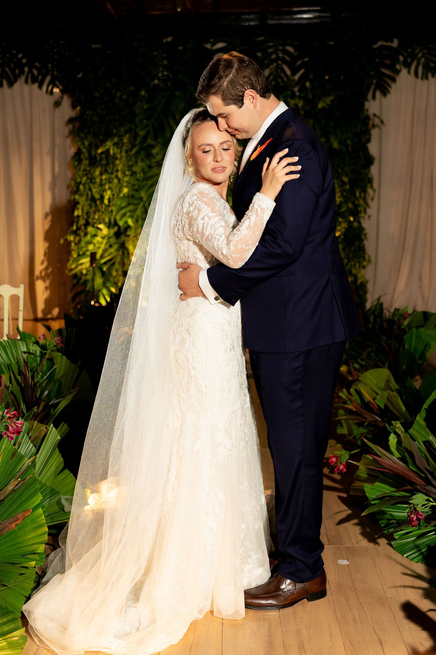 Bride and groom share intimate first dance embrace, she in lace gown with veil, he in navy suit, surrounded by tropical greenery.