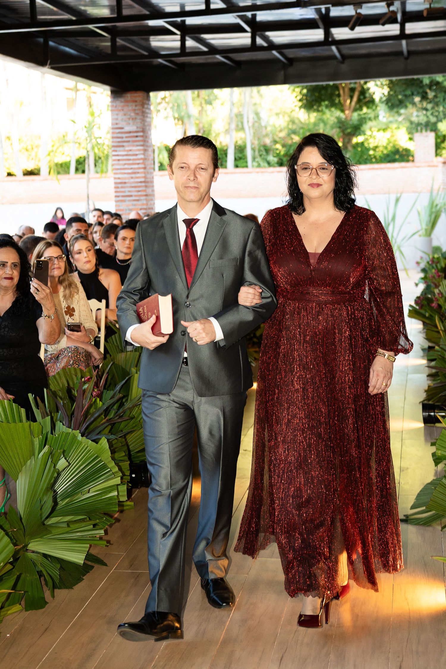 Elegant couple in matching burgundy outfits walking down aisle at outdoor wedding ceremony.
