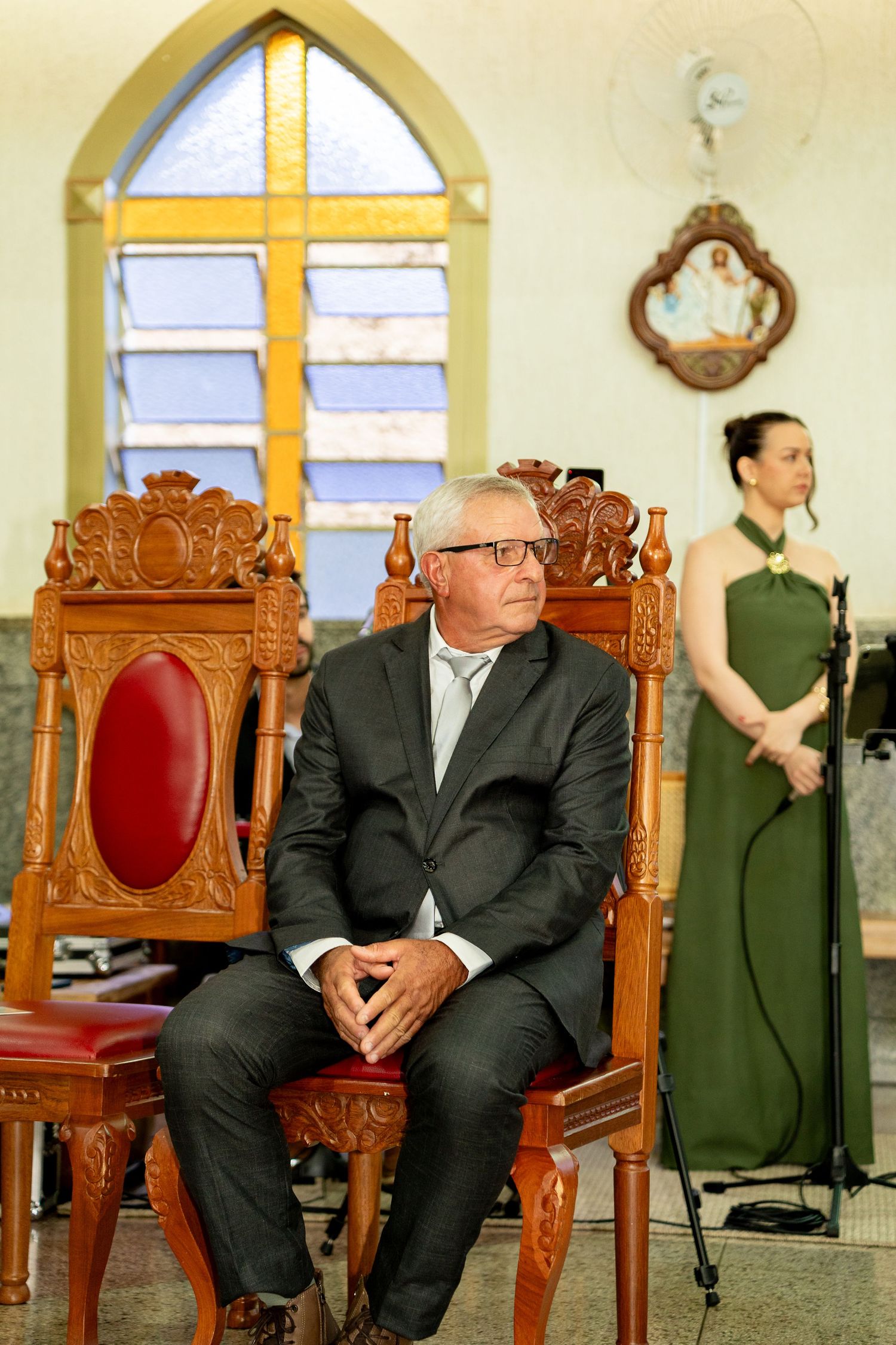 Elderly man in suit sits on ornate wooden chair in church with stained glass cross window and woman in green dress.