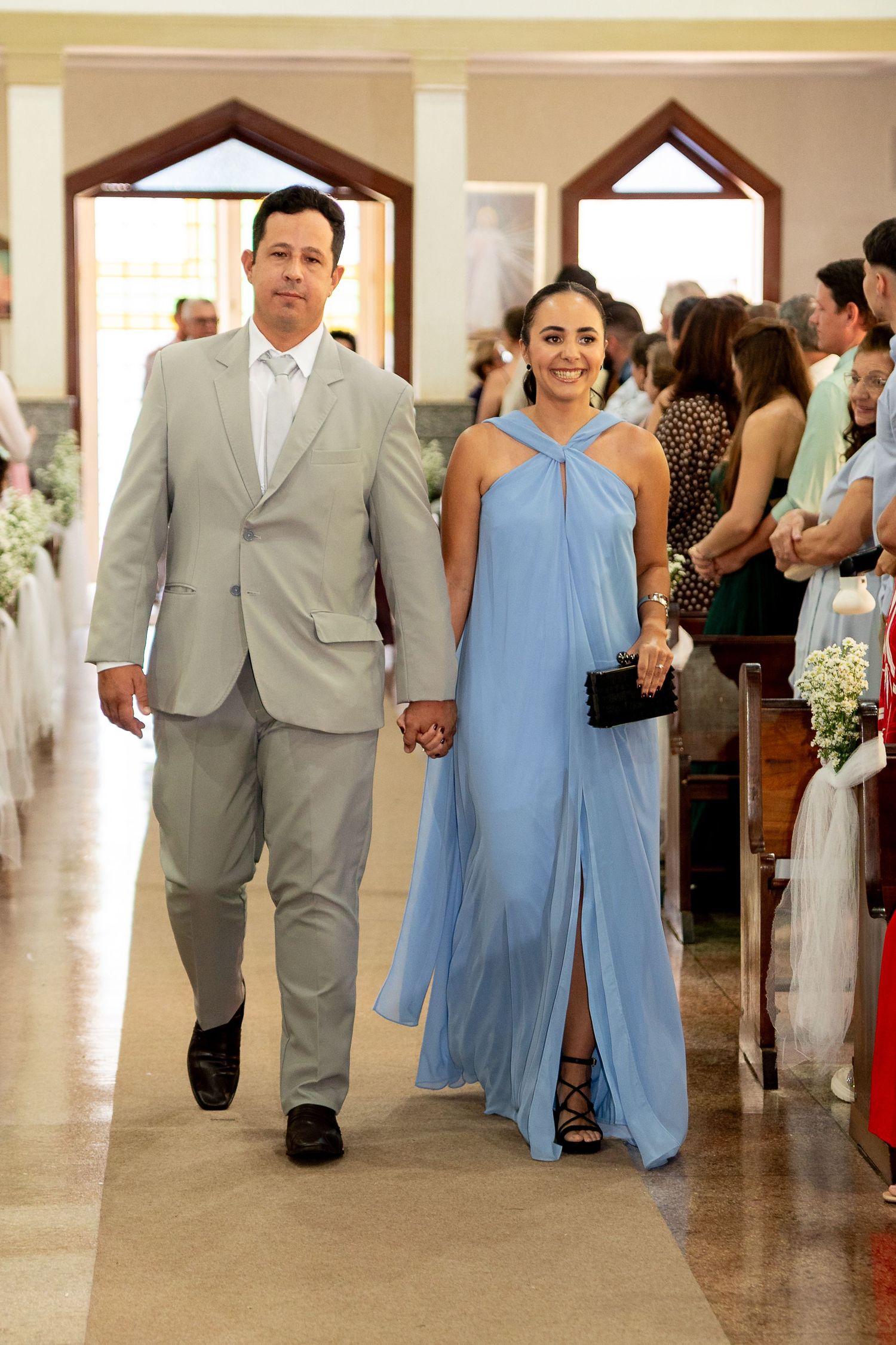 A couple walks hand in hand down a church aisle, man in gray suit, woman in light blue halter gown.