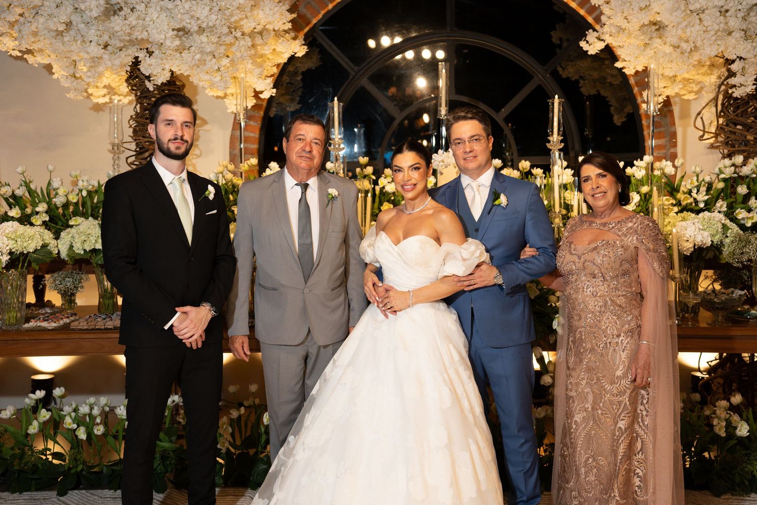 Elegant wedding ceremony with bride in white gown and groom in blue suit posing with family amid floral decor.