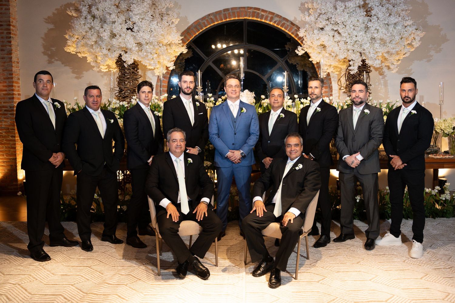 Groom in blue suit poses with groomsmen in black tuxedos at elegant wedding venue with floral arch backdrop.