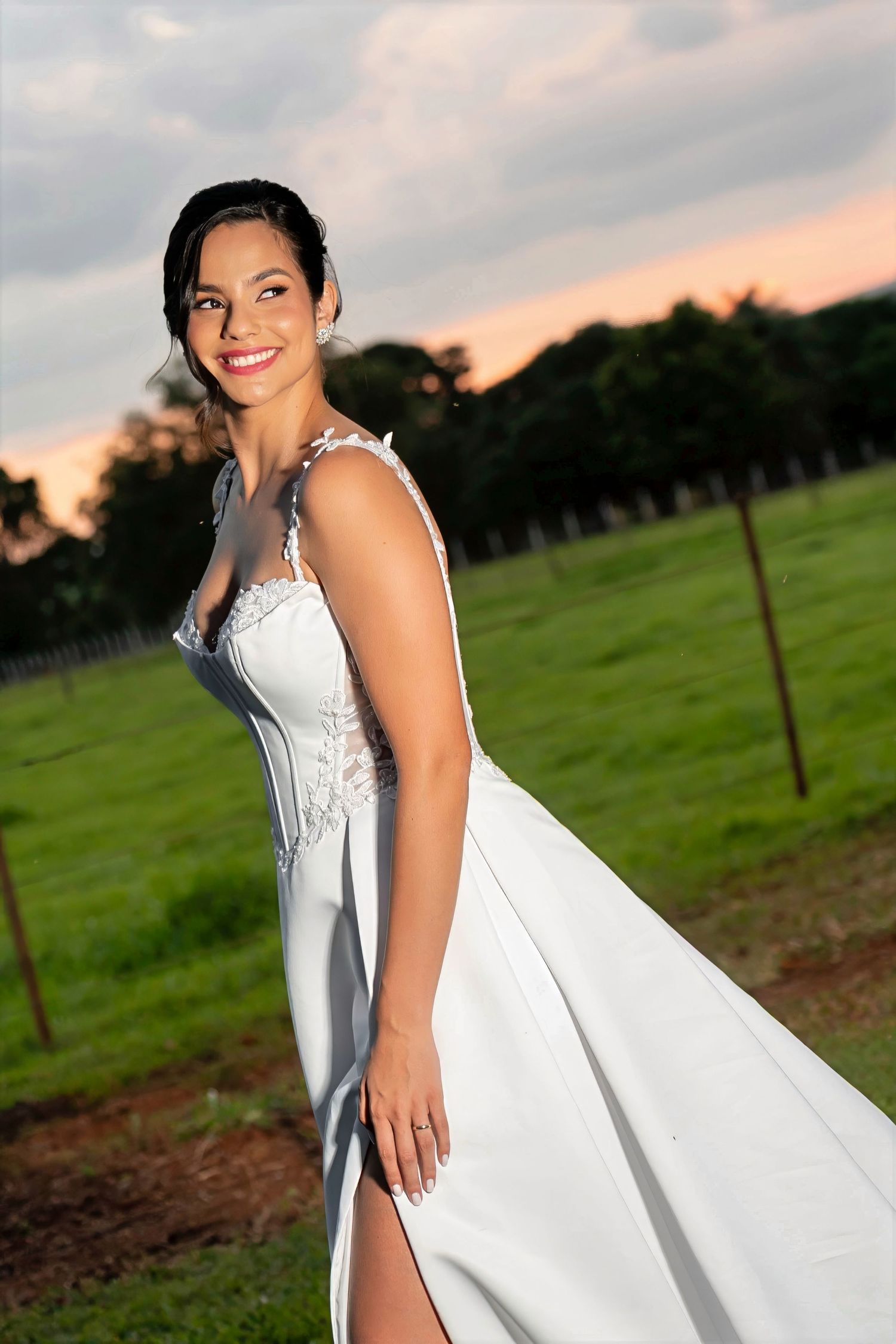 Smiling bride in white lace wedding gown poses outdoors at sunset on green field.