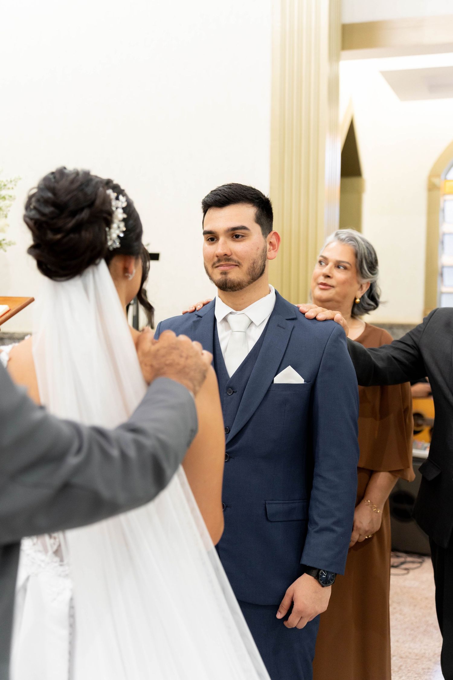 Groom in navy suit stands at church altar facing bride in white dress and veil during wedding ceremony.