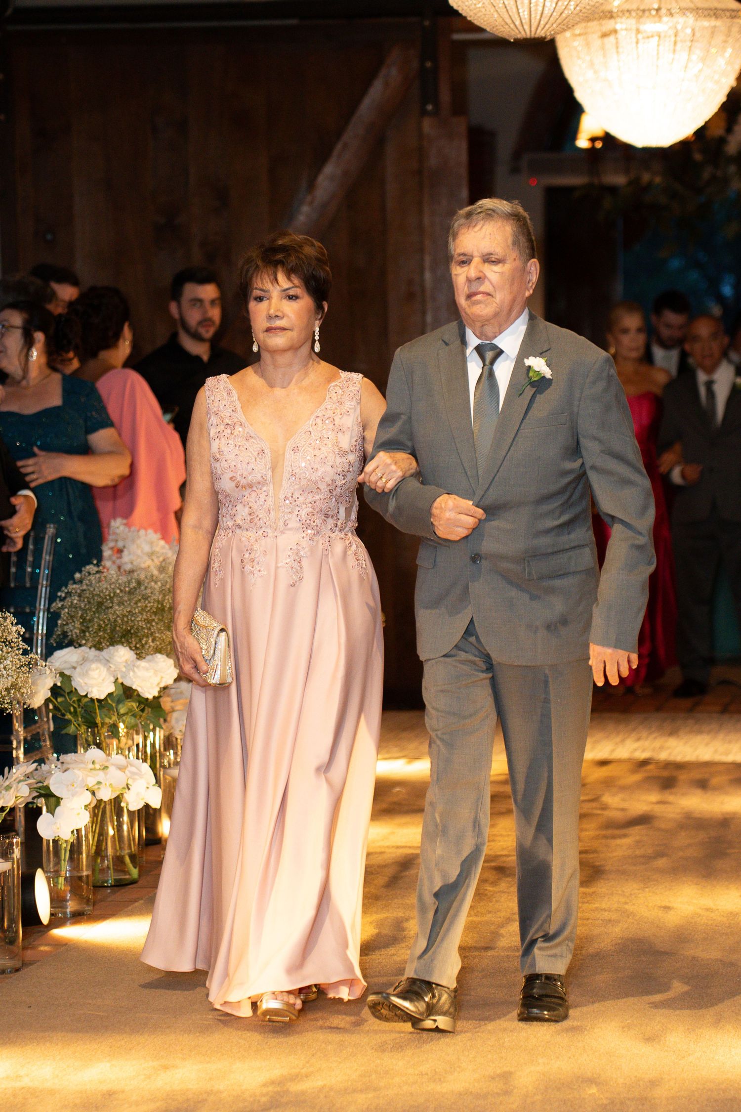 Elegant couple walking down wedding aisle, woman in pink gown, man in gray suit with boutonniere, rustic venue.