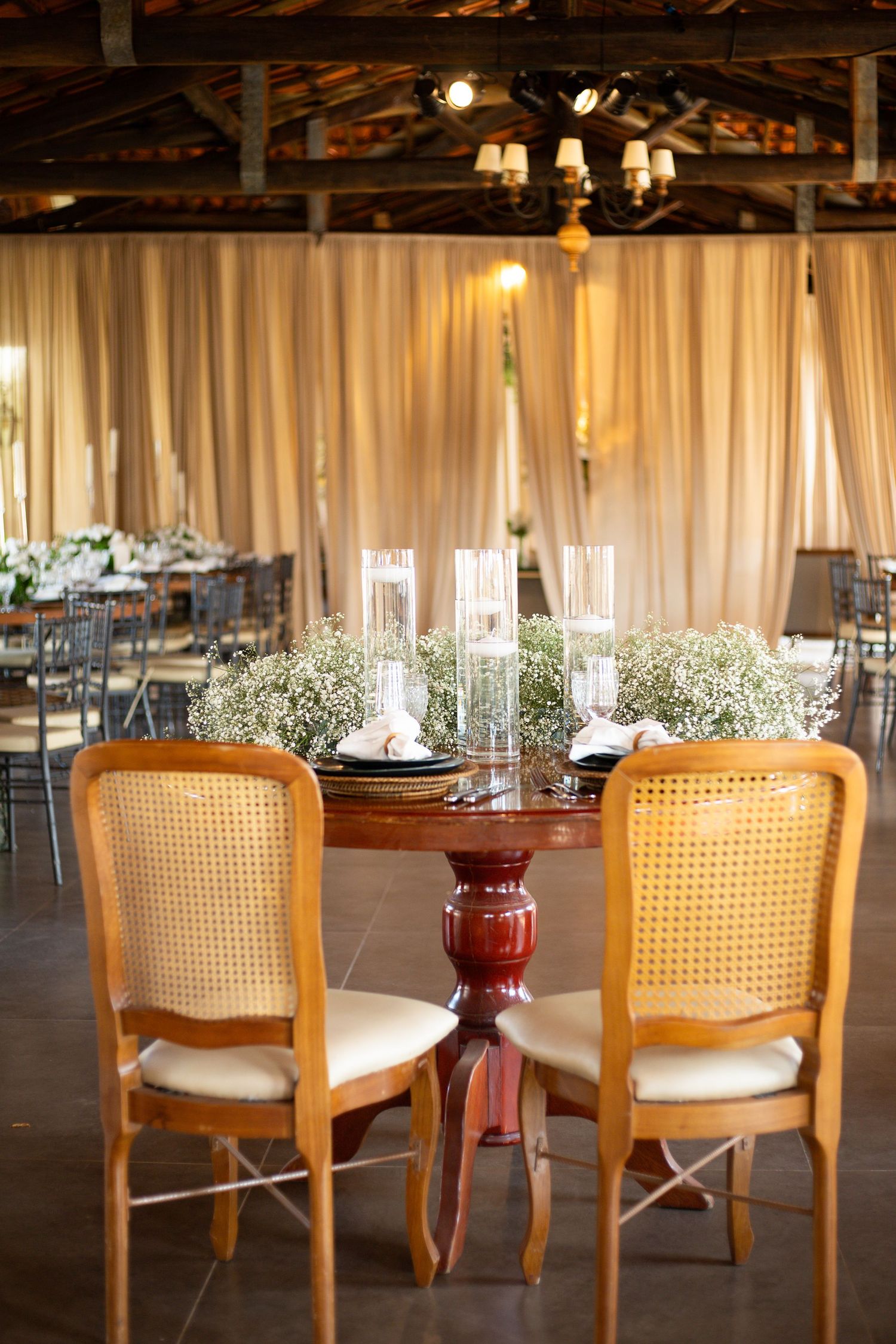 Elegant wedding reception table with wicker chairs, baby's breath centerpiece, and candles in a draped barn venue.