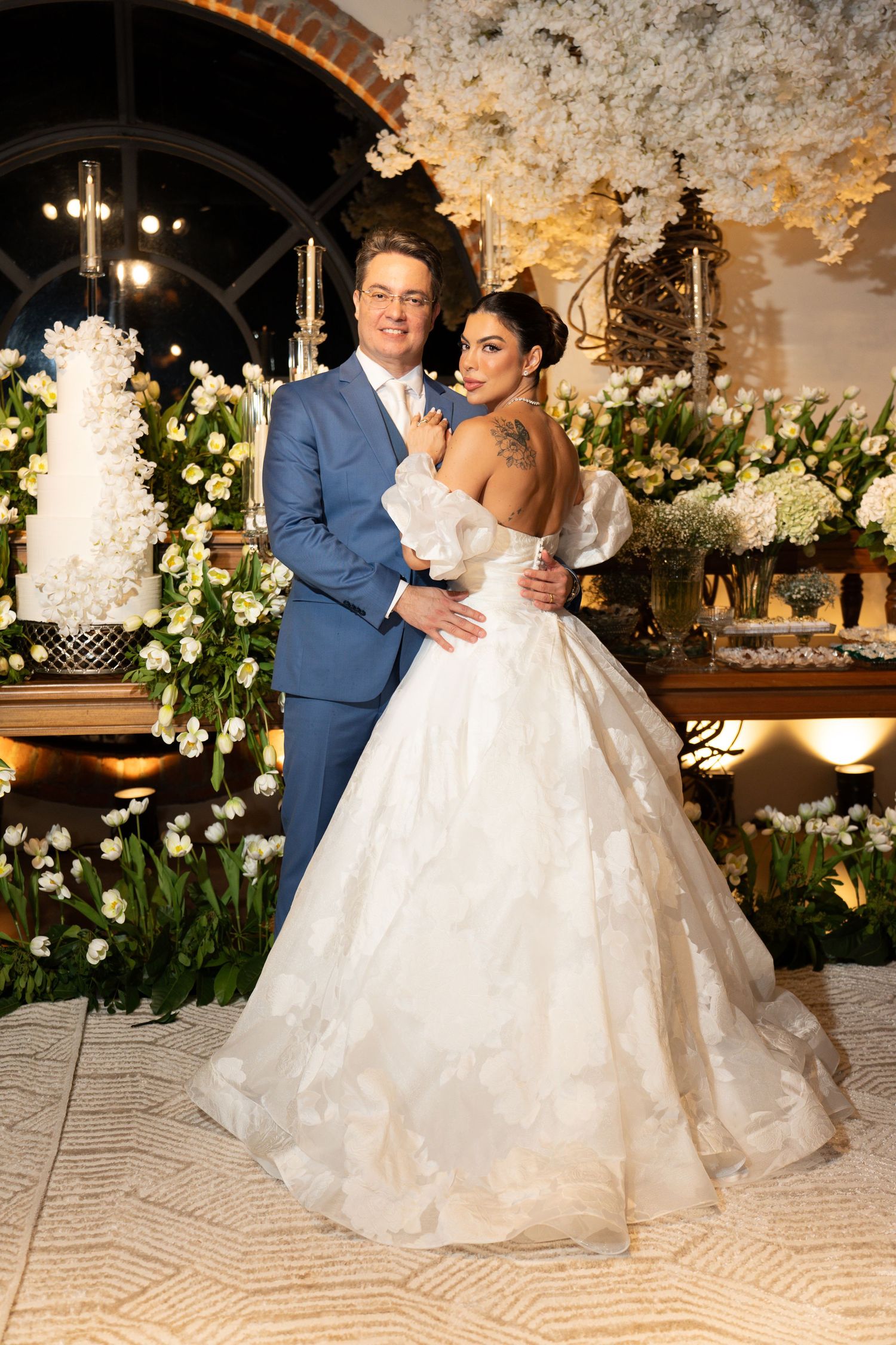 Elegant bride in a white ballgown and groom in blue suit pose together at a floral wedding reception.