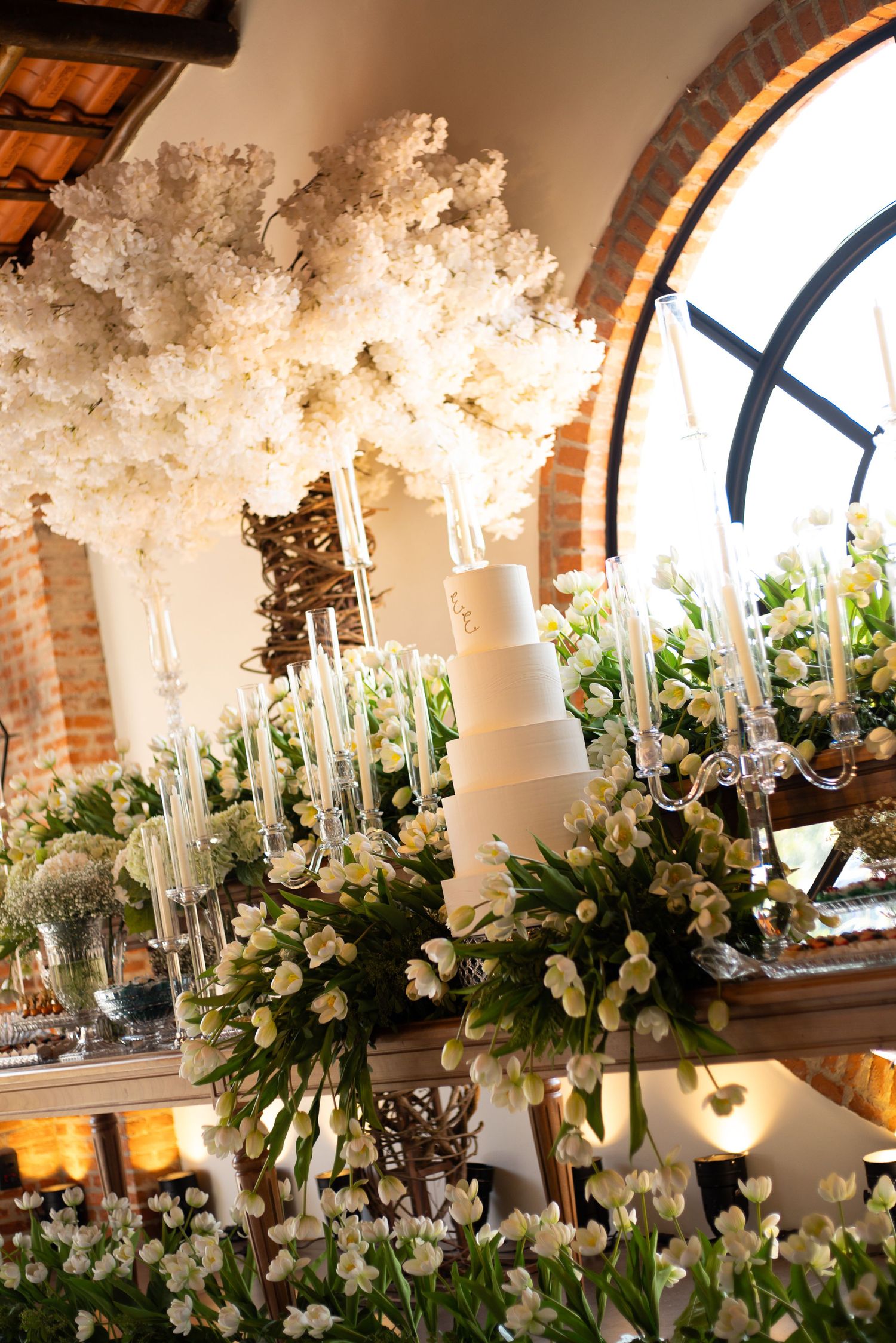 Elegant wedding cake display with white floral arrangements, candelabras, and a large clock window in a rustic venue.