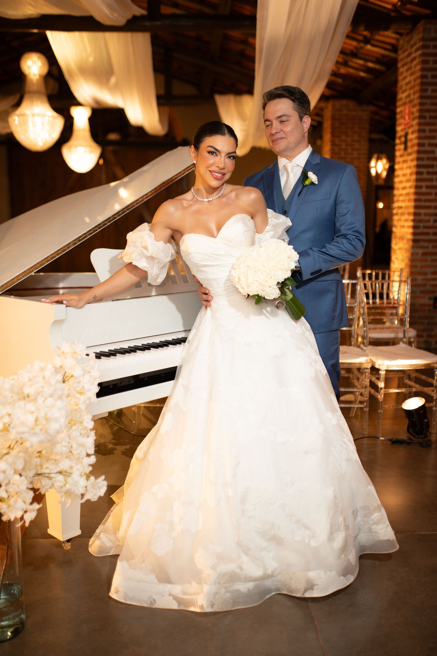 Bride in white ball gown holds bouquet beside white grand piano as groom in blue suit stands beside her.