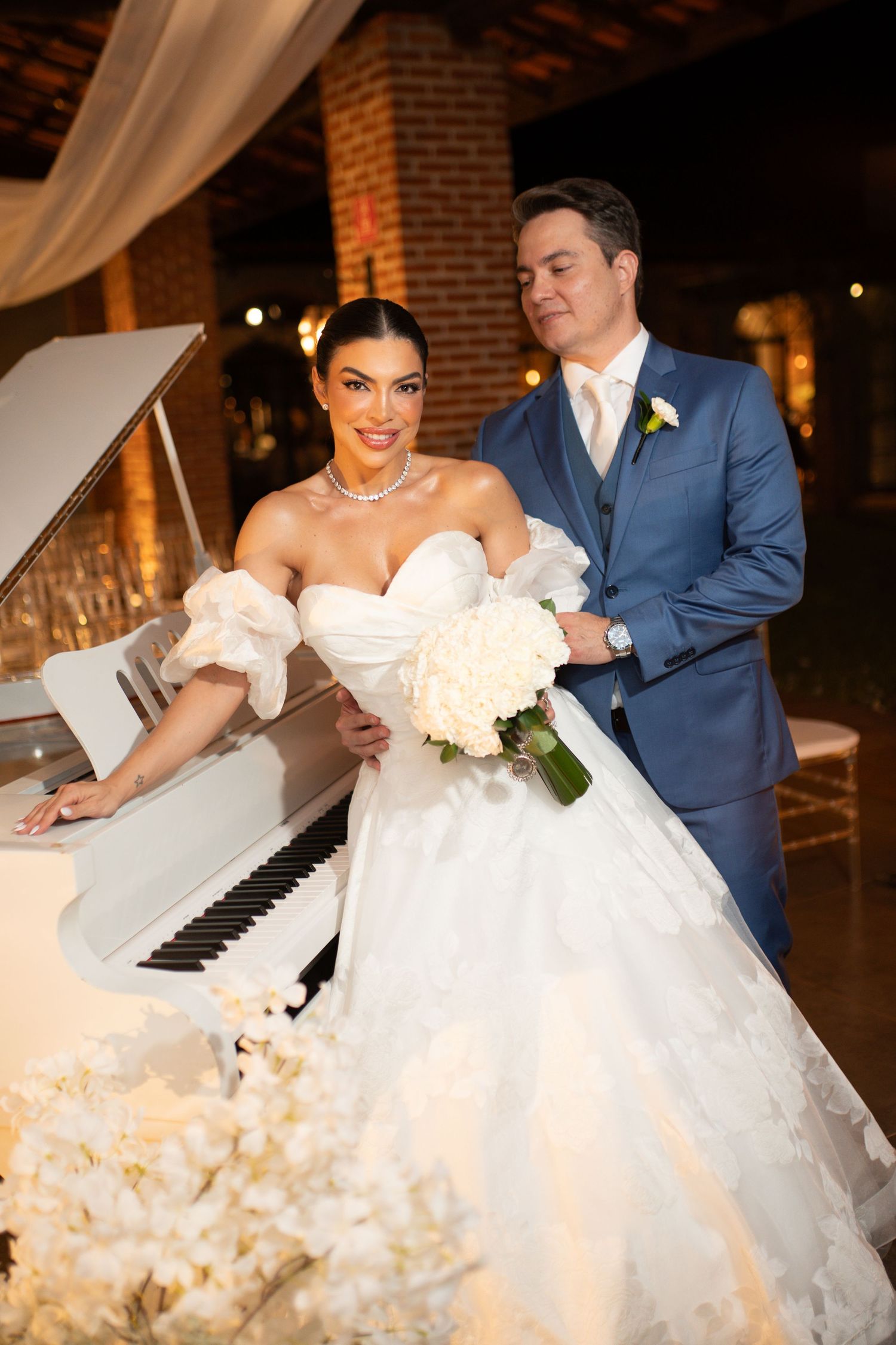 Bride in white ballgown with bouquet poses beside white grand piano with groom in blue suit at elegant wedding venue.