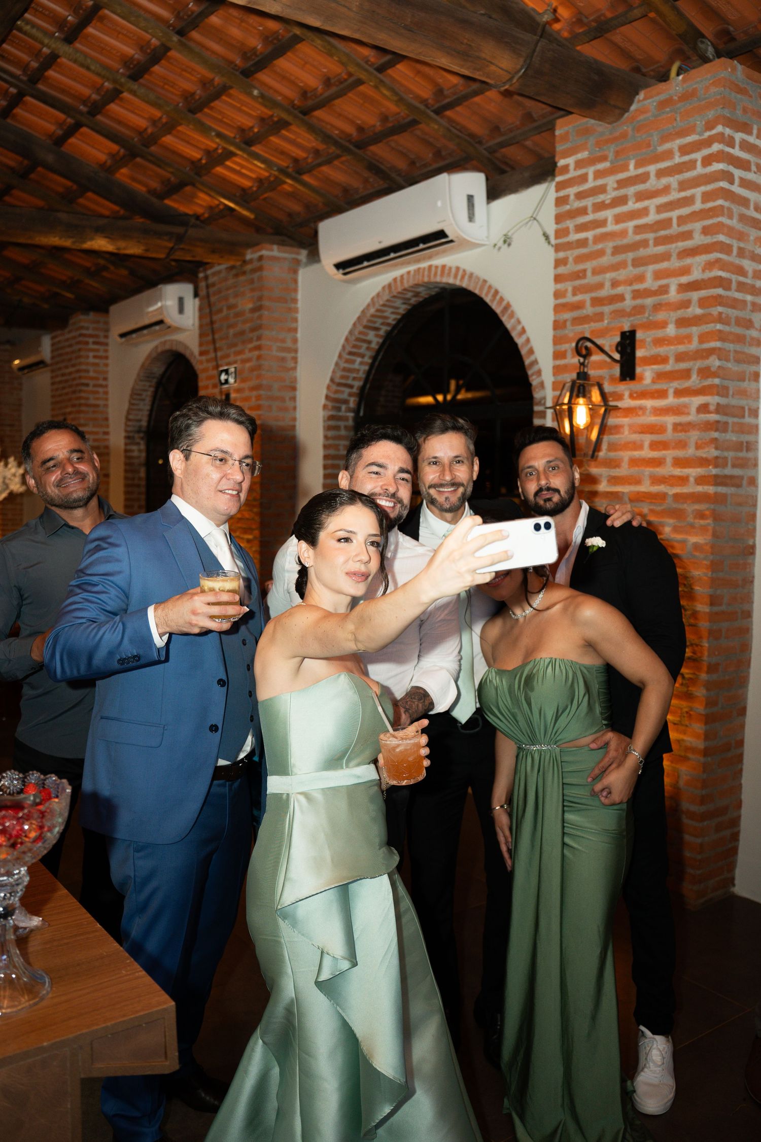 Wedding guests in sage green dresses take a group selfie at an elegant brick venue reception.