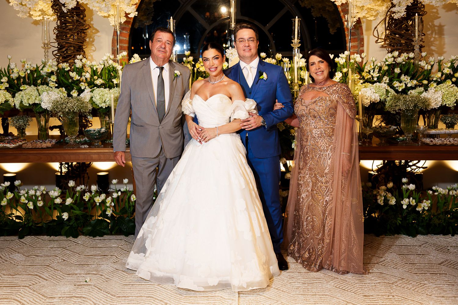 Elegant wedding photo with bride in white gown, groom in blue suit, and family members in formal attire at a floral venue.