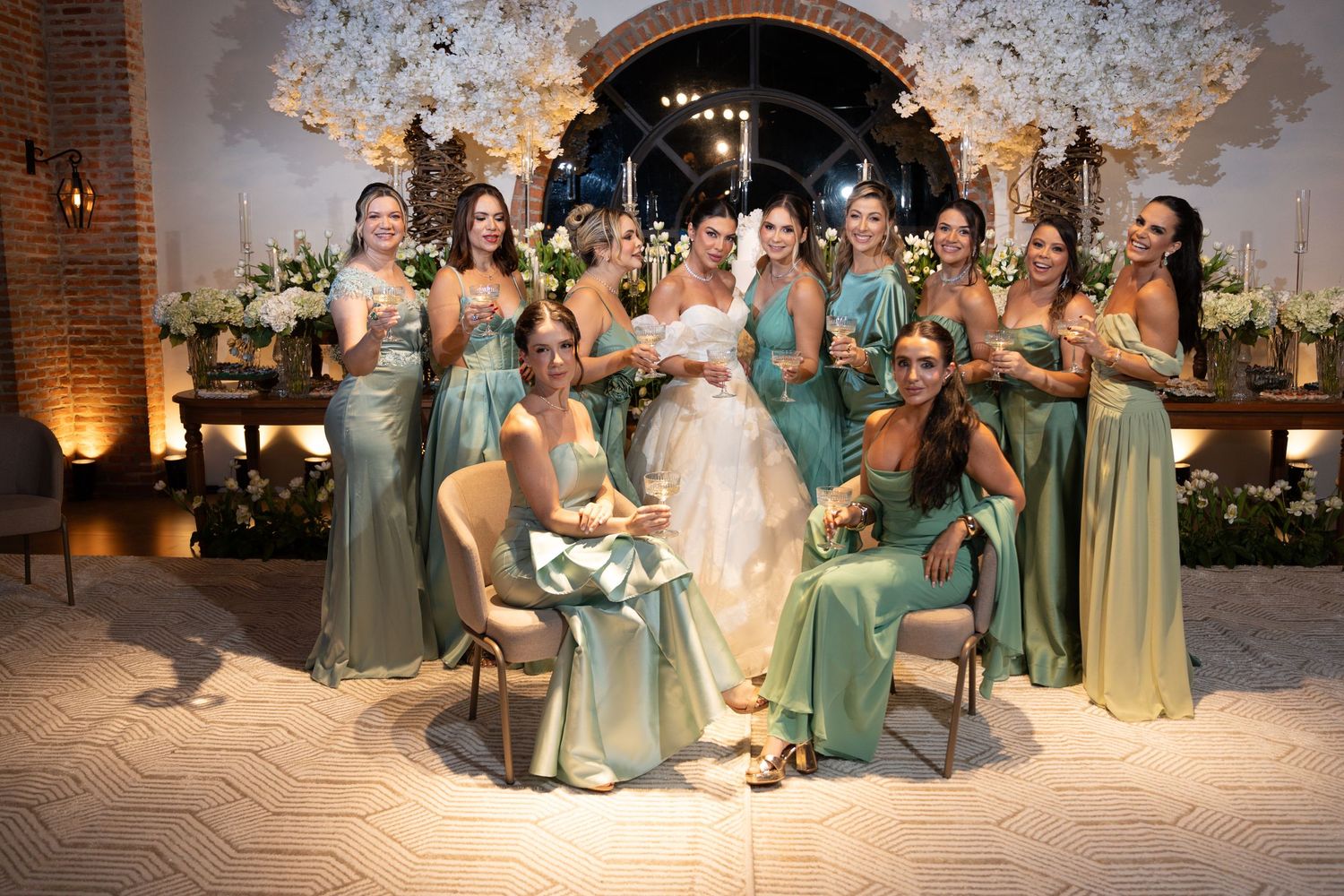 Bride in white gown poses with bridesmaids in sage green dresses at elegant floral wedding venue.