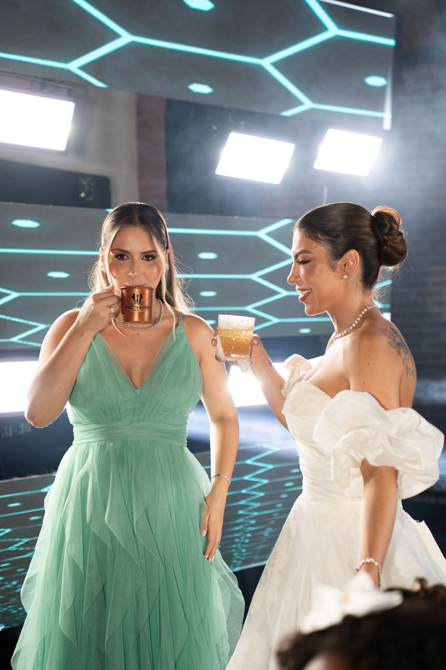 Two elegant women in formal dresses toasting with beer mugs at a glamorous event with hexagonal neon lights.