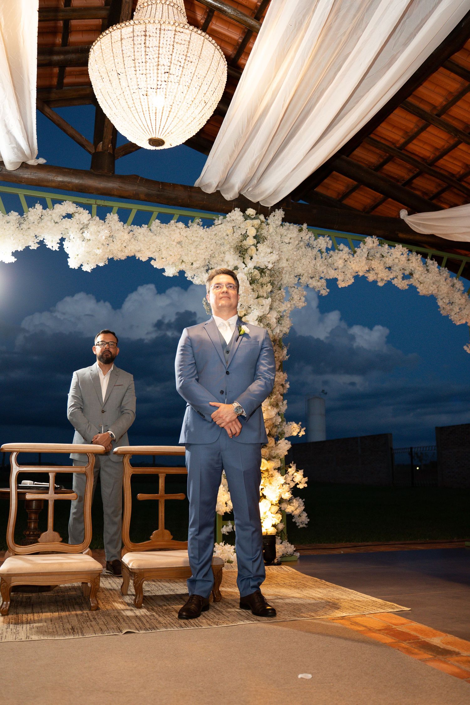 Groom in blue suit stands at outdoor wedding altar with floral arch and chandelier at dusk.