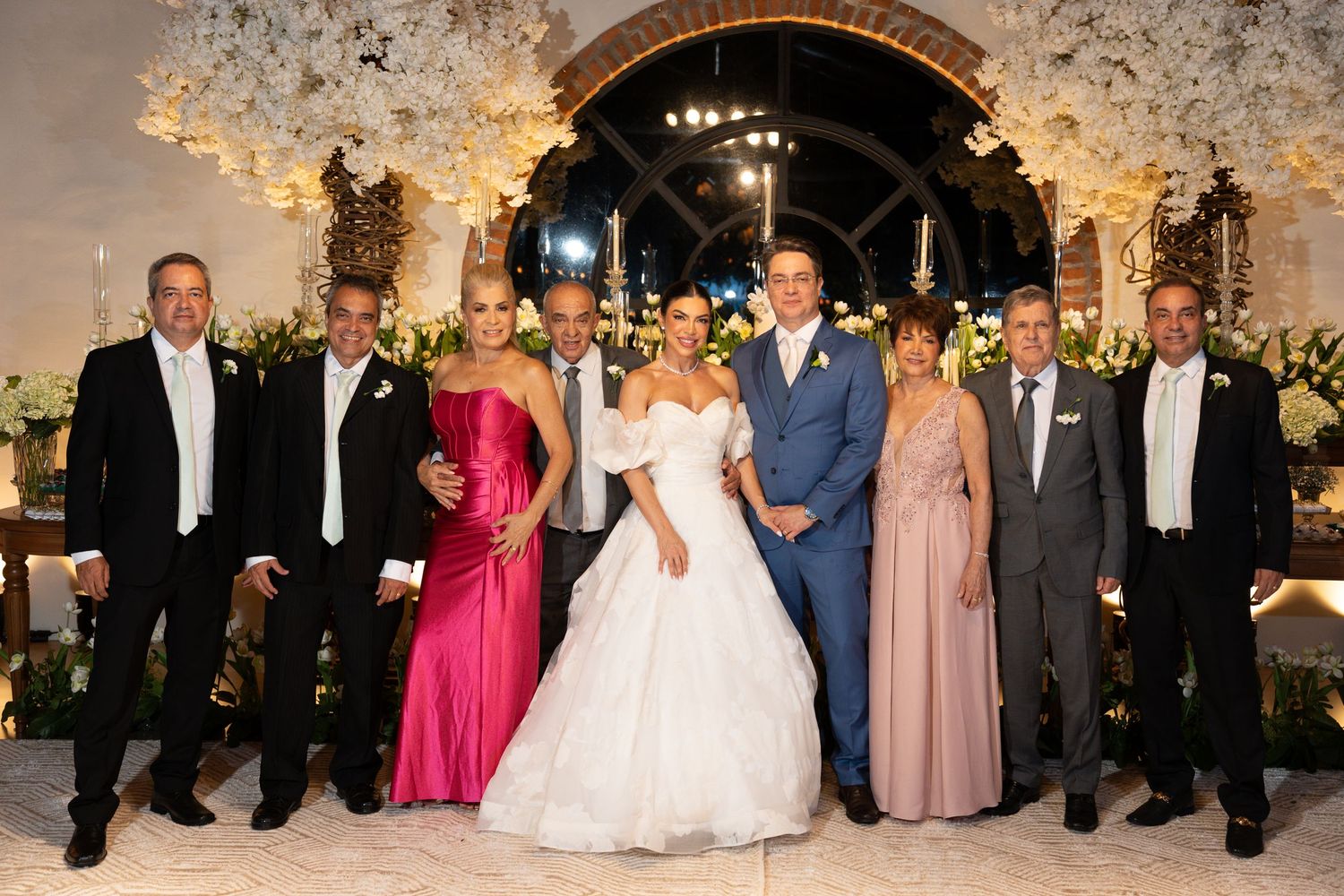 Wedding party poses elegantly in formal attire before floral arch, bride in white gown with groom and family members.