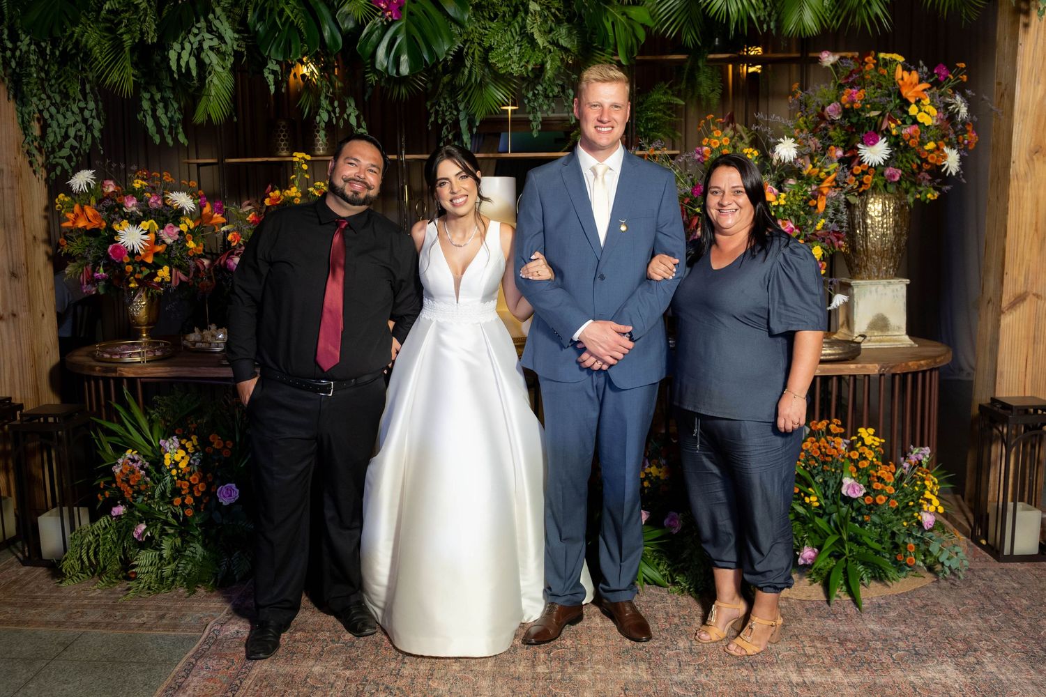 Bride in white gown and groom in blue suit pose with two guests in front of colorful floral backdrop.