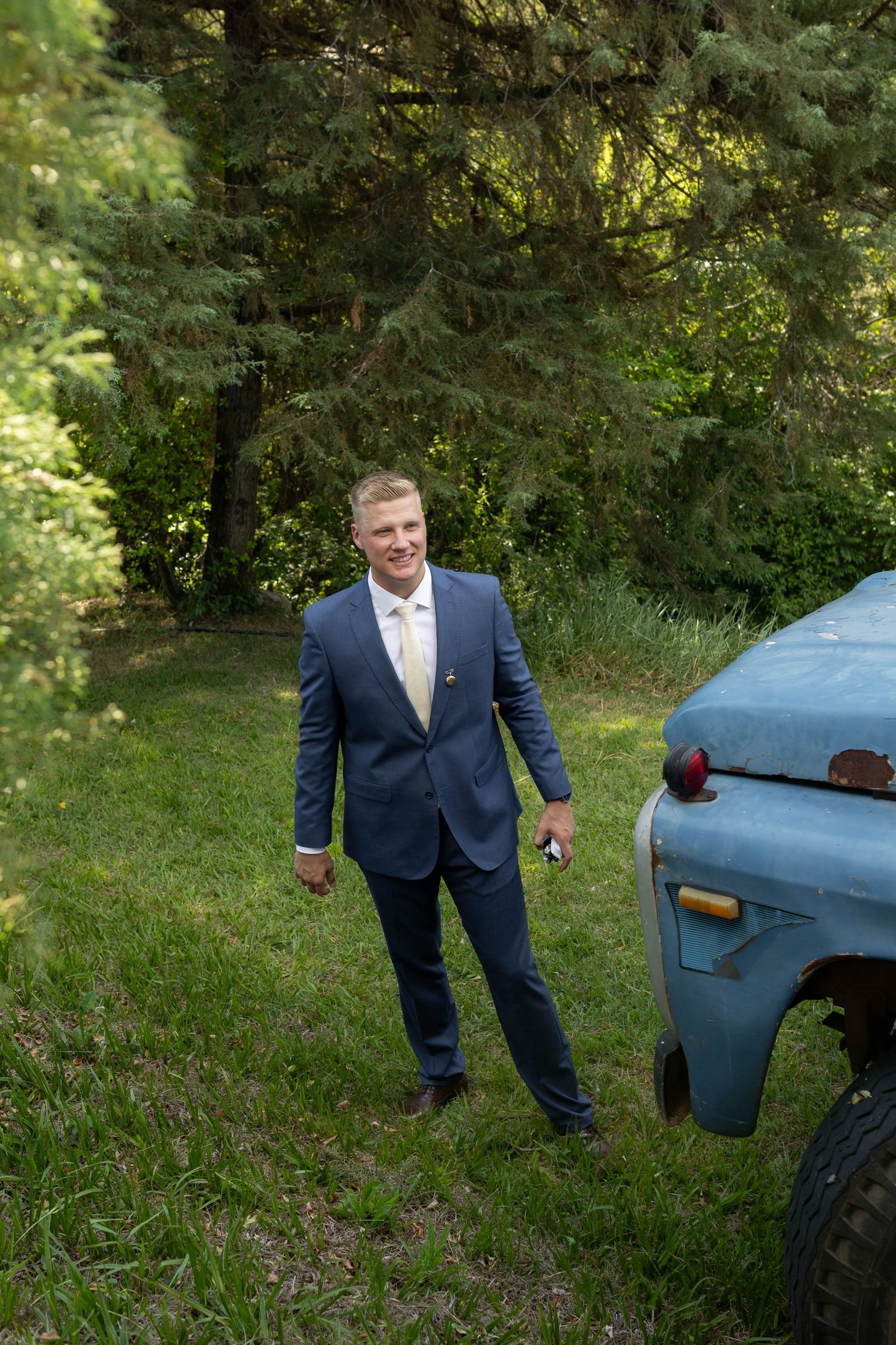 A smiling groom in a navy suit and tie stands on green grass beside a vintage blue truck surrounded by lush trees.