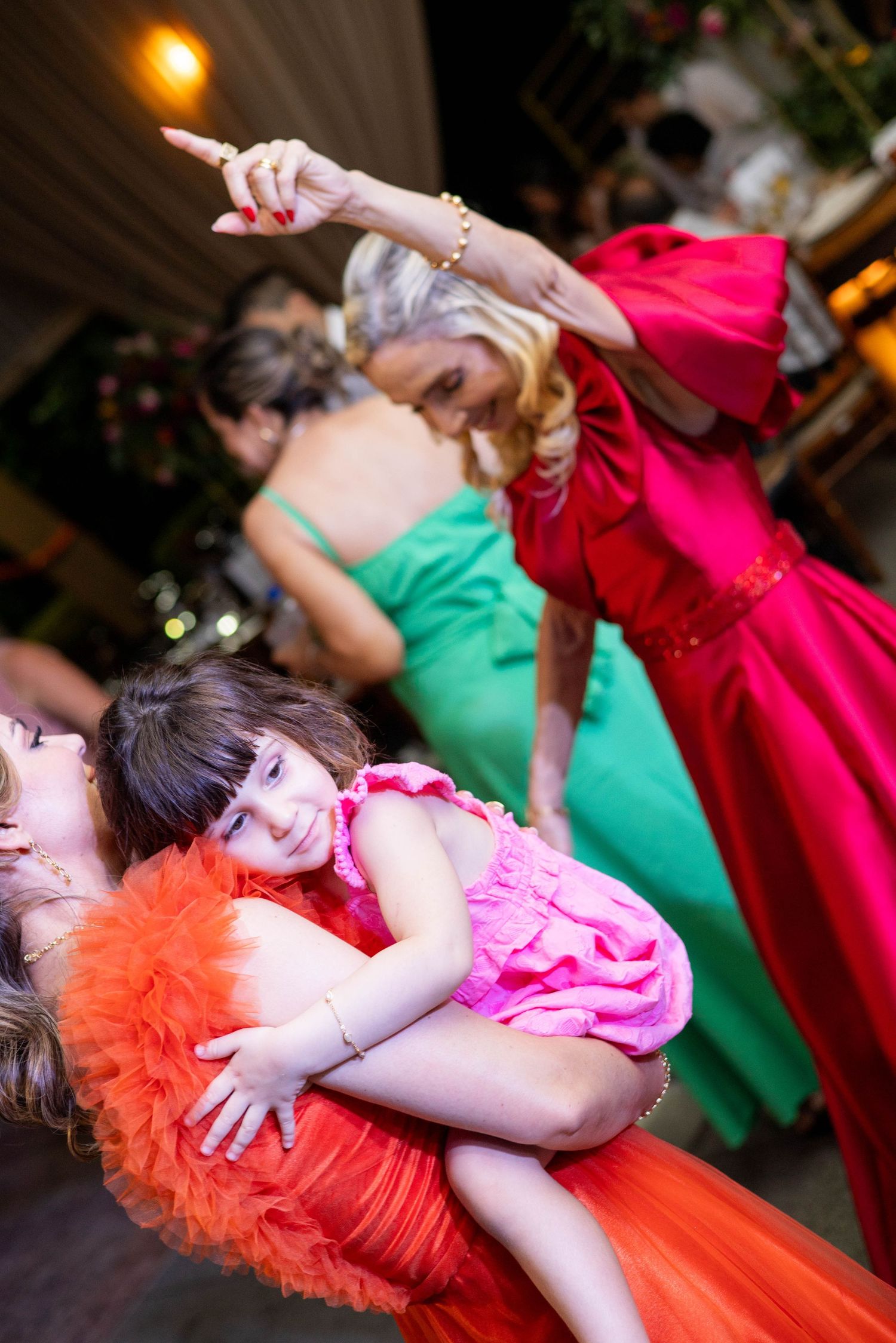 A young girl in pink dress being held at a festive event with women in colorful gowns dancing in background.