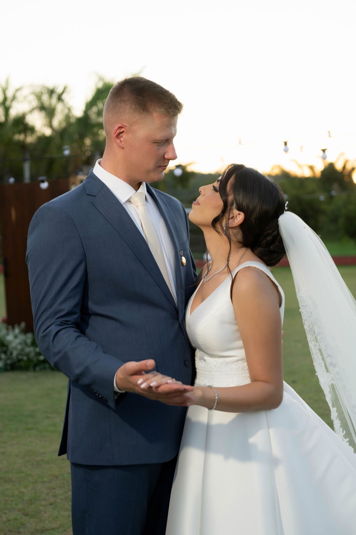 Bride and groom share an intimate moment outdoors at golden hour, she in a white gown with veil, he in a navy suit.