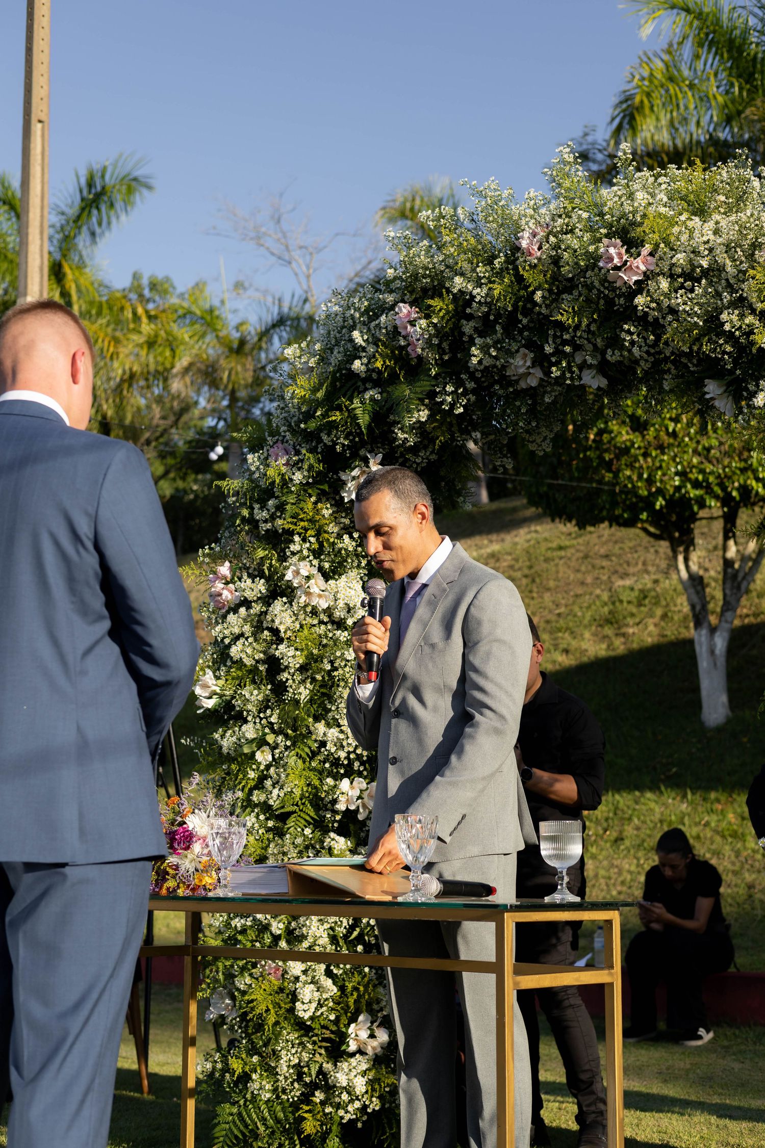 Officiant in gray suit speaks at outdoor wedding ceremony with floral arch, lush greenery, and blue sky backdrop.
