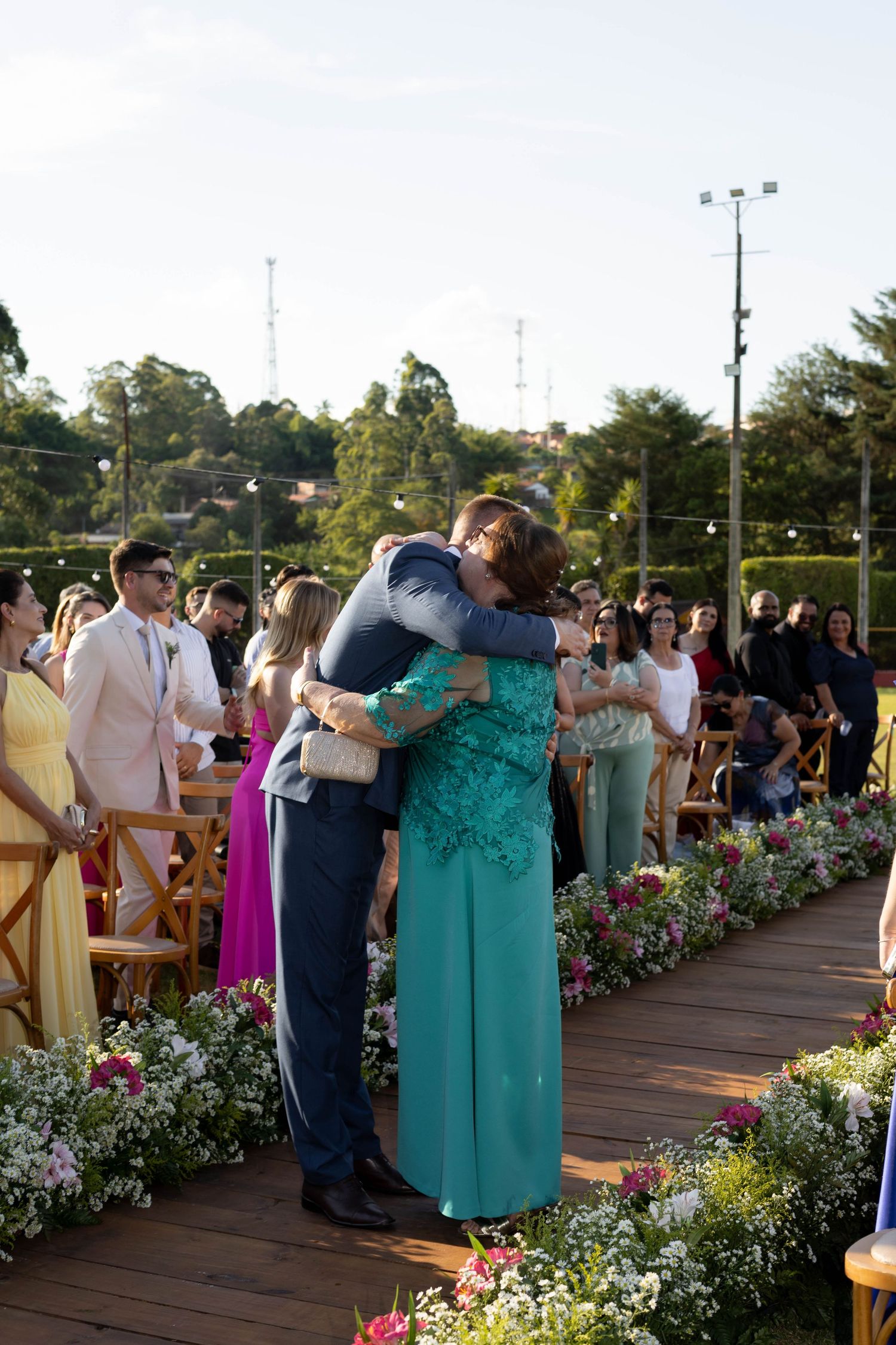 Groom hugging a woman in teal dress at outdoor wedding ceremony with floral aisle decorations.