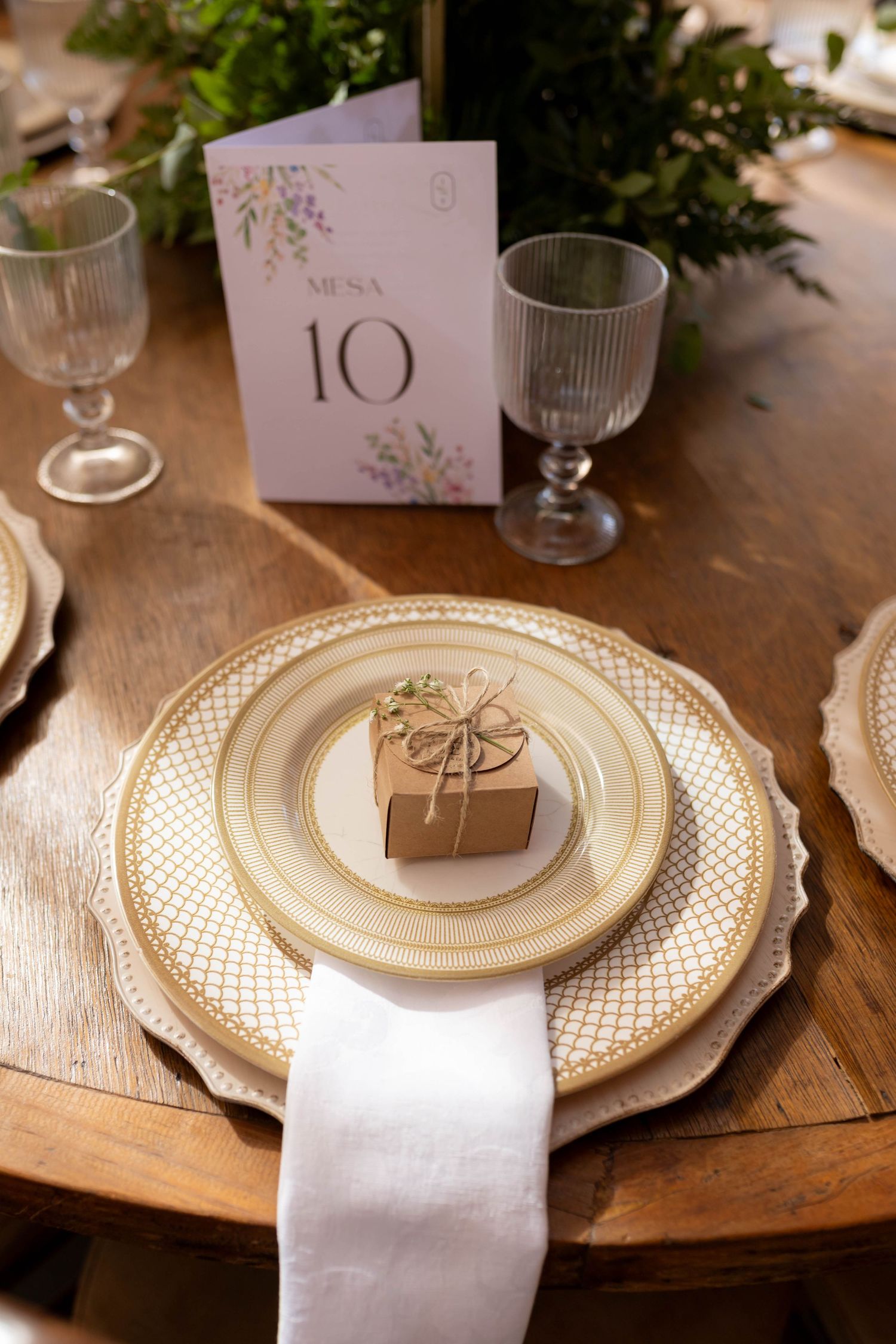 Elegant wedding table setting with gold-rimmed plates, kraft favor box, wine glasses, and a floral table number 10 card.
