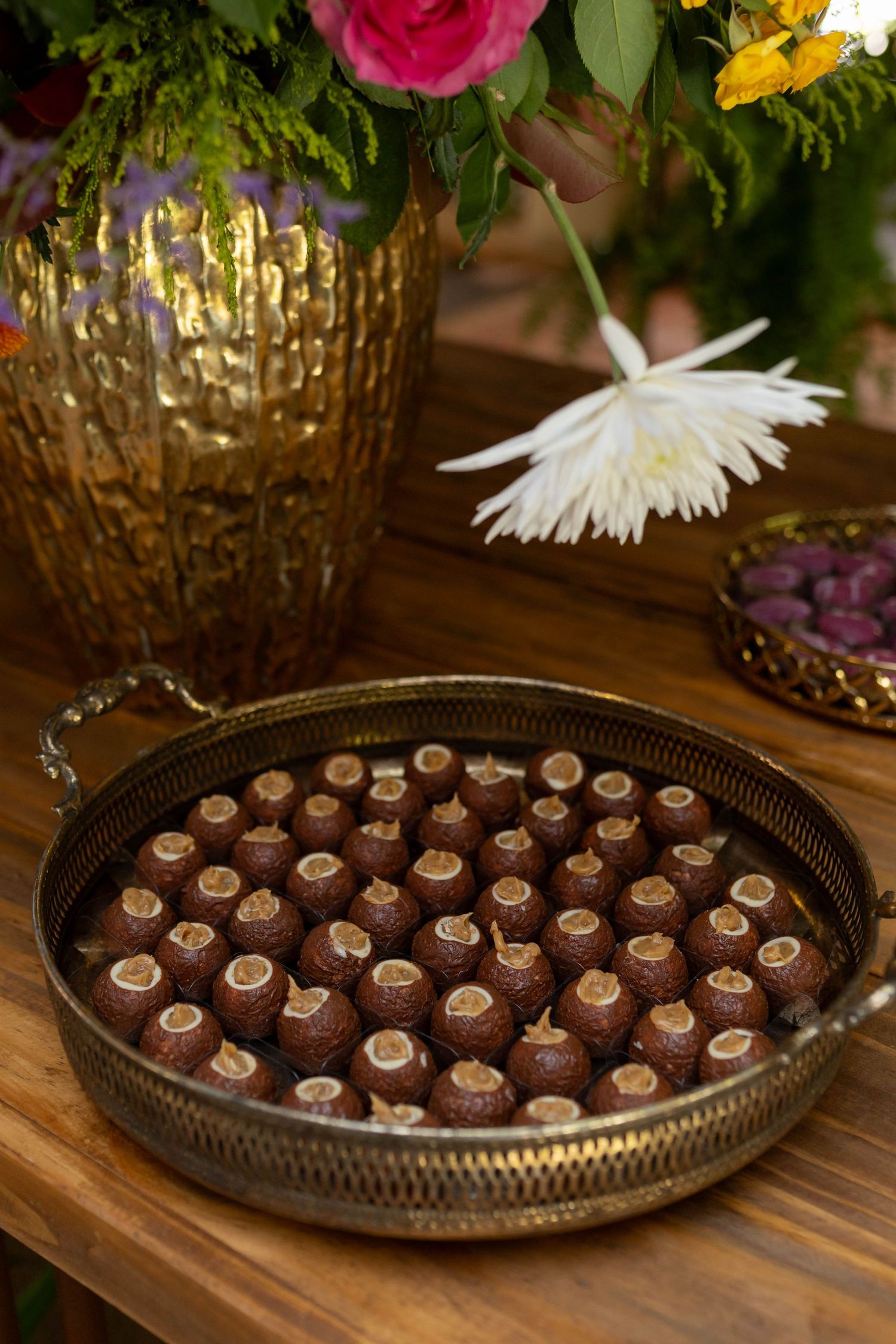 Gold tray filled with chocolate truffles on wooden table, surrounded by colorful floral arrangements in golden vase.