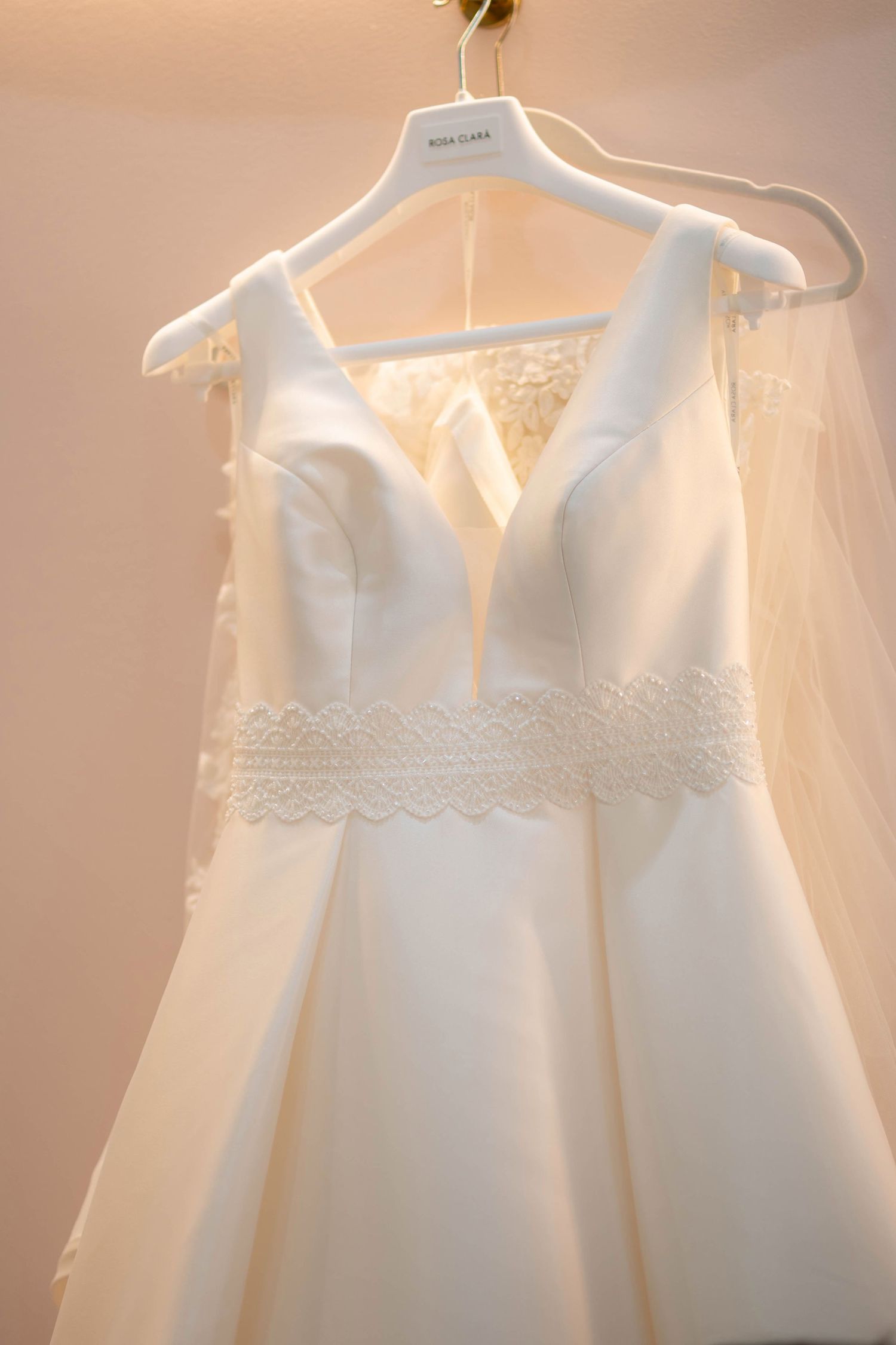 Elegant Rosa Clara white wedding dress with lace waist detail and V-neckline displayed on a hanger with veil.