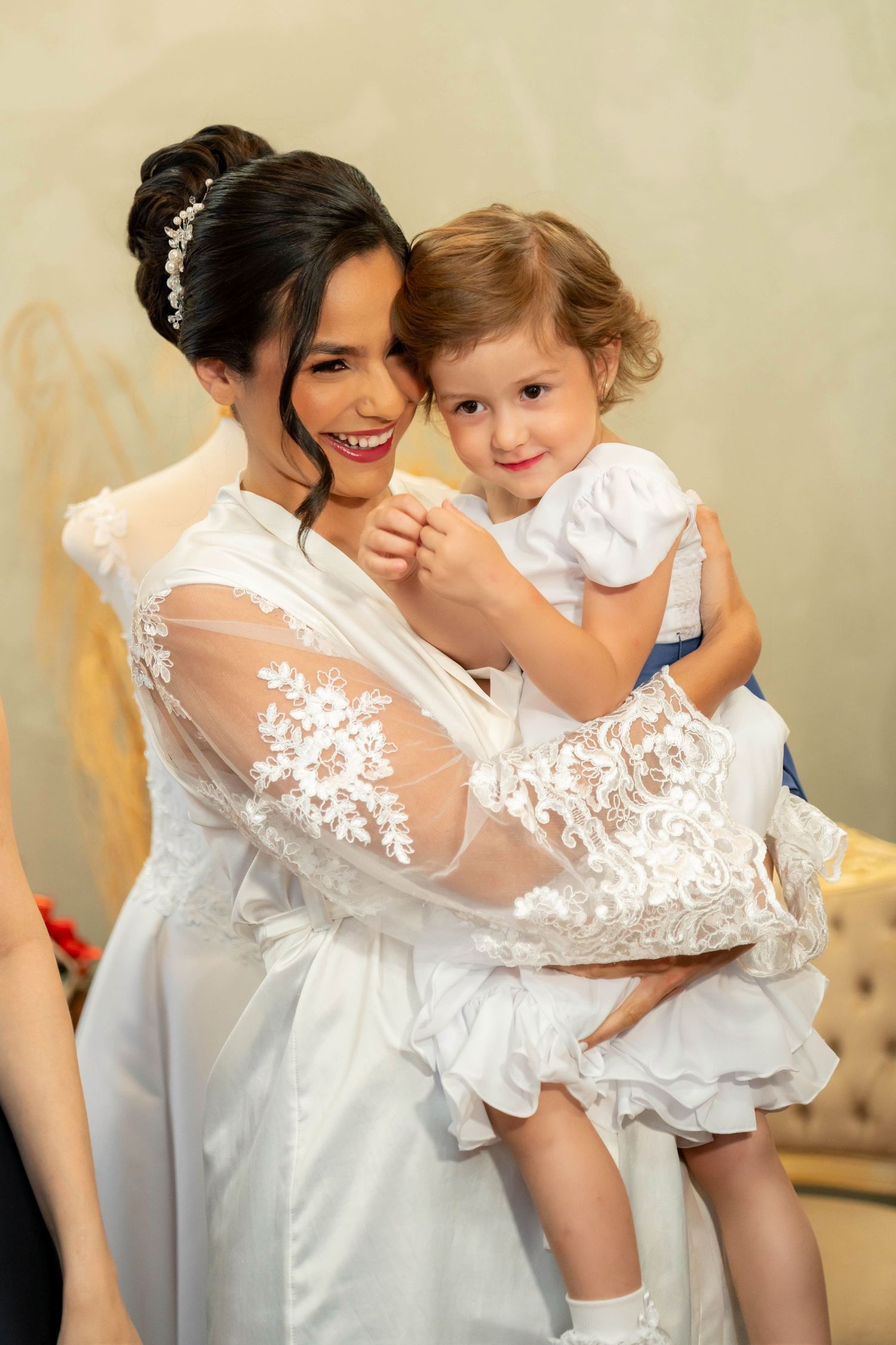 Smiling bride in white lace robe holds a young girl in a white dress during wedding preparations.