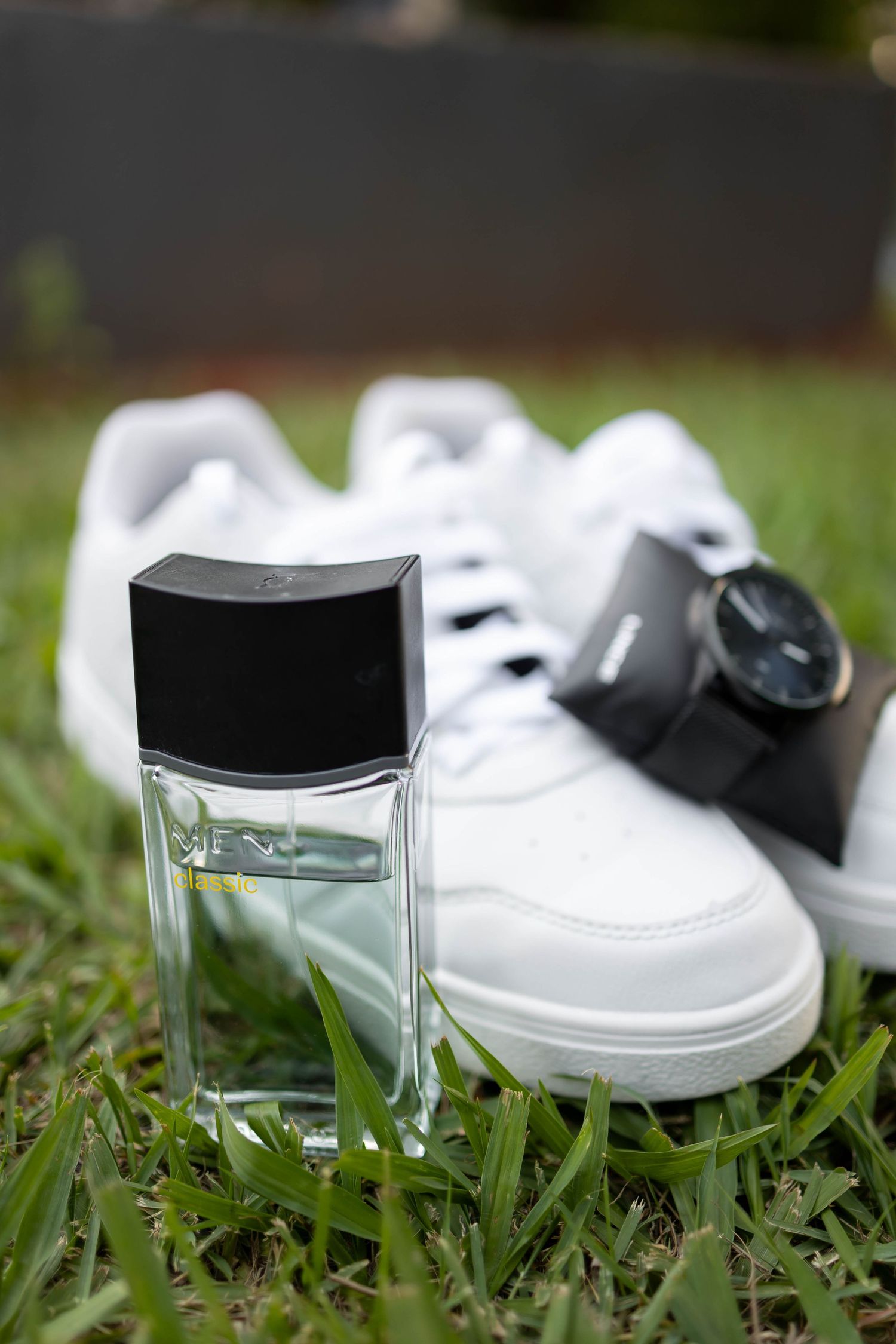 Glass cologne bottle with black cap on grass beside white sneakers and a dark watch outdoors.